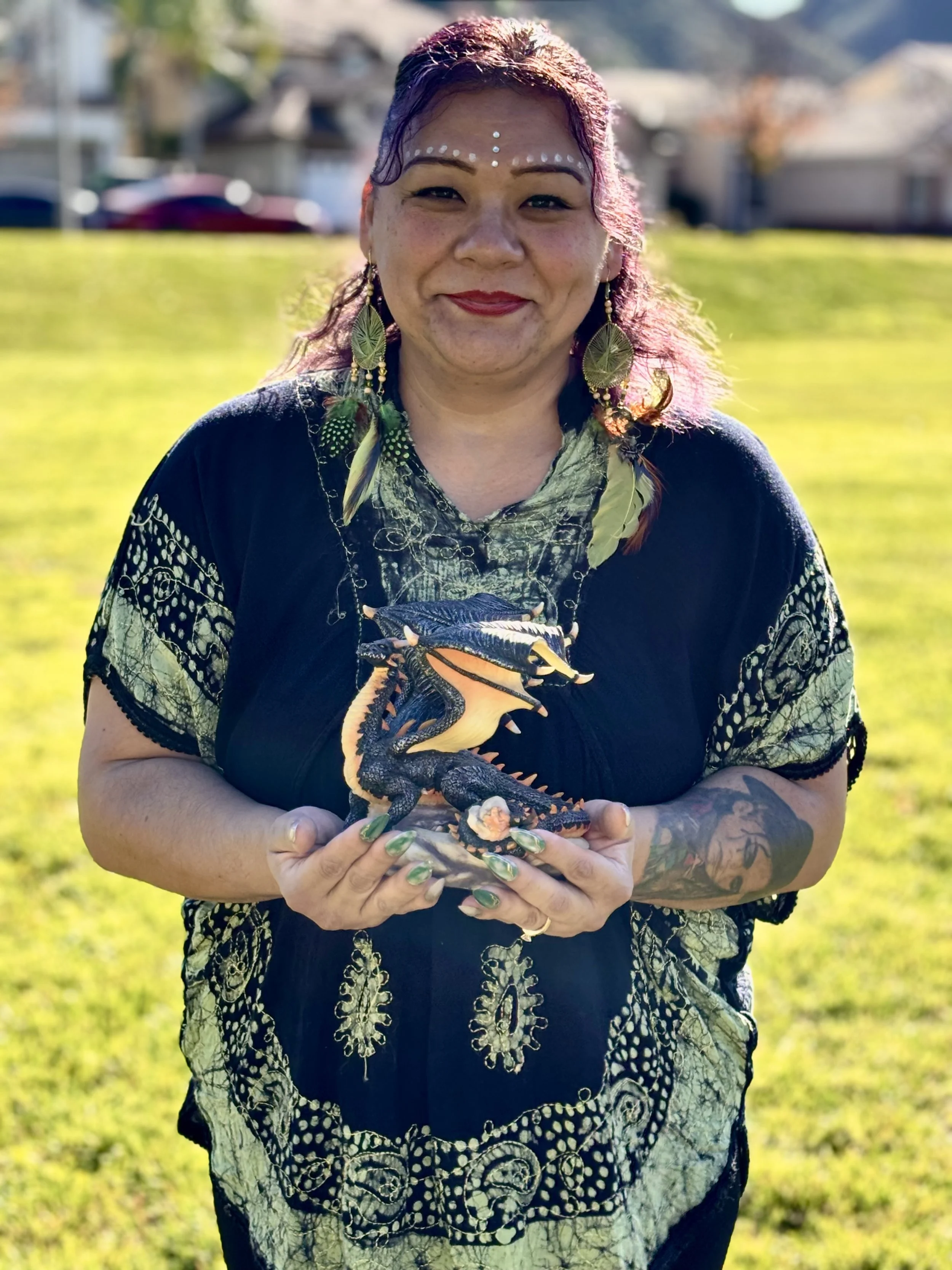 A woman with purple hair and tattoos holding a small dragon figurine outdoors on a sunny day.