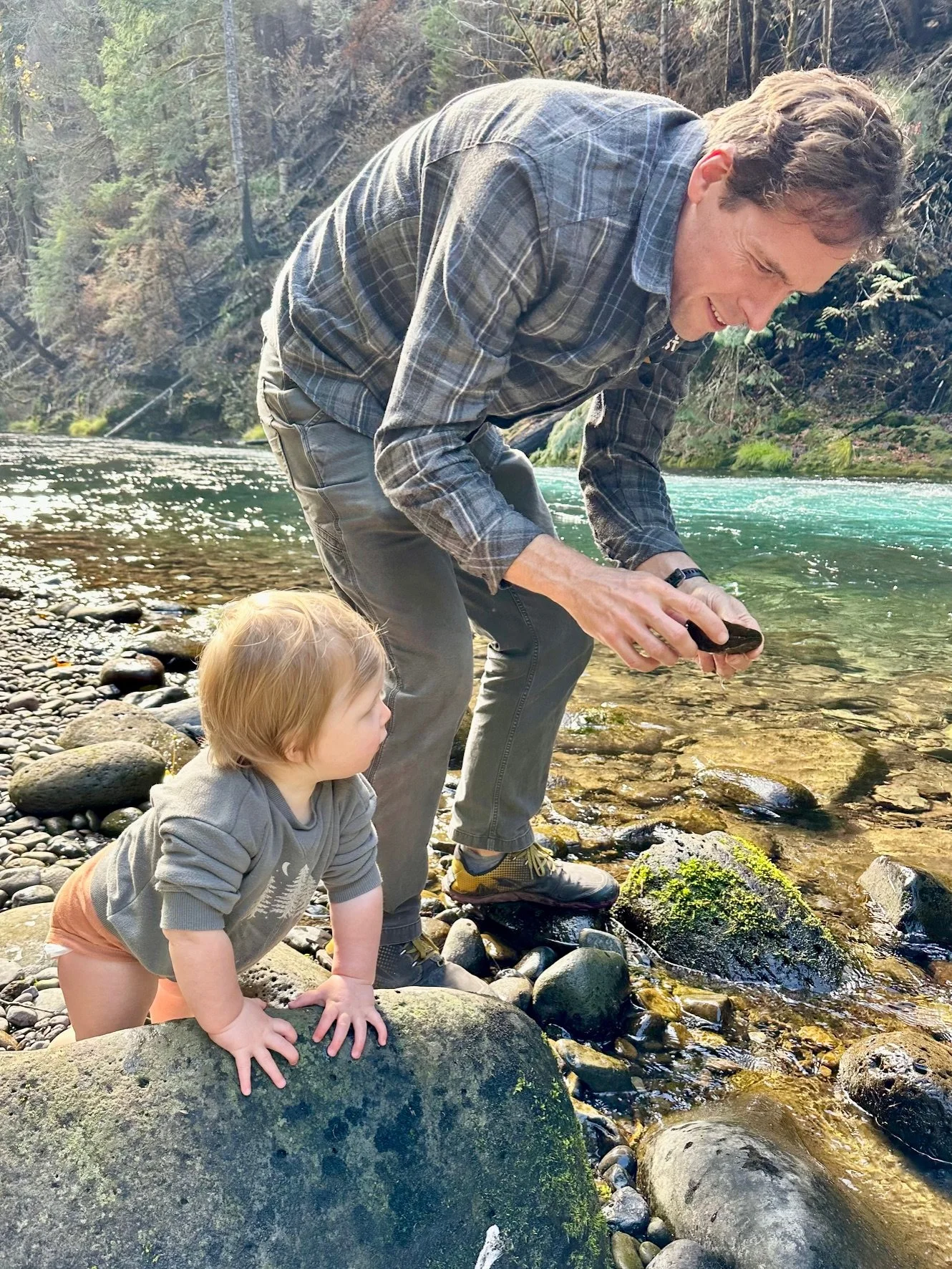 A man and a young child by a river. The man is squatting, holding a small object, and wearing a plaid shirt and hiking shoes. The child, with light hair, is crawling on a large rock, watching the man. The river is clear, with a forested area in the background.