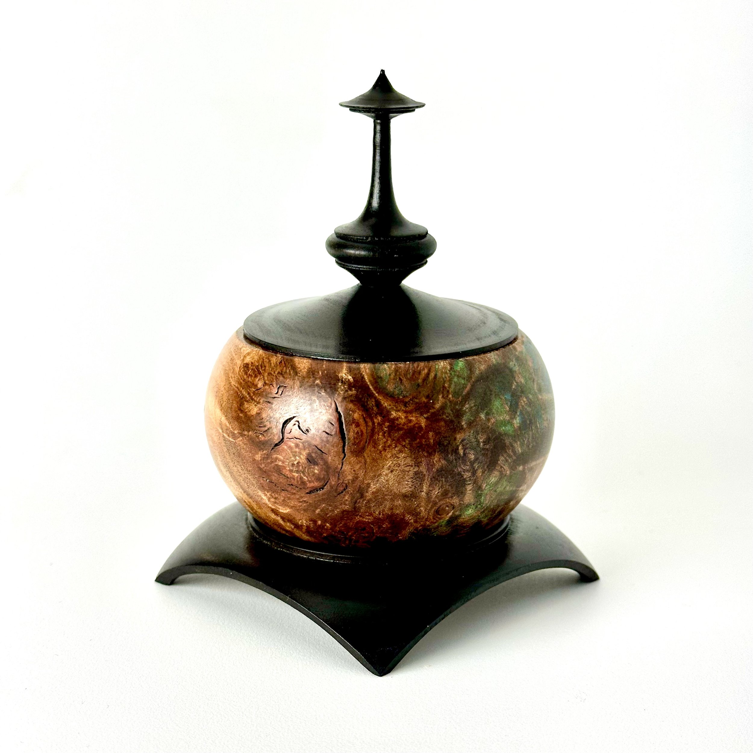 A decorative container with a round, wooden-textured body, topped with a black lid and a ornate black finial, placed on a black curved stand.