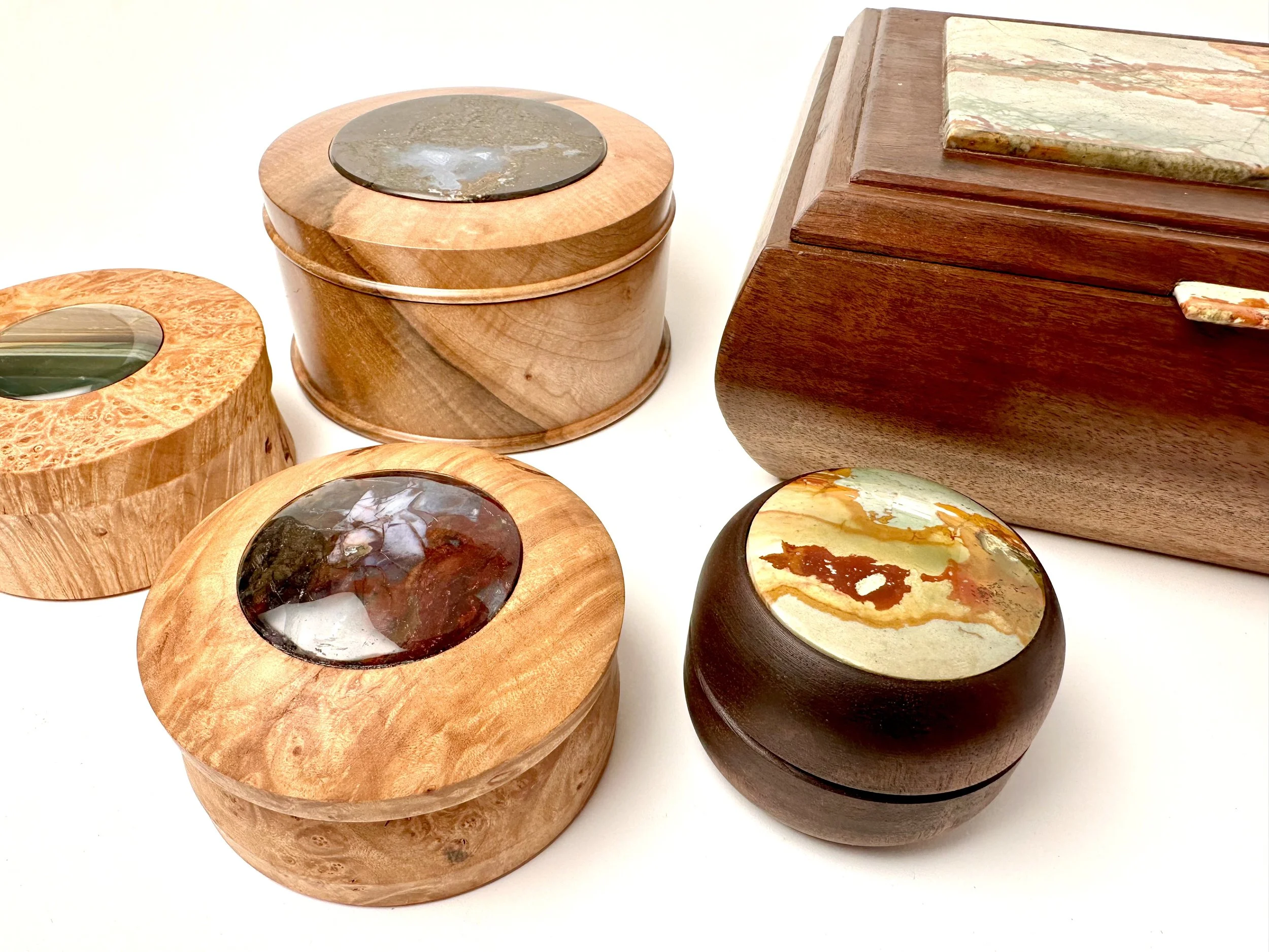 Various handcrafted wooden boxes made fro maple burl and walnut with colorful stone and gemstone inlays including Owyhee picture jasper and Blue Mountain Jasper and Purple Cow Jasper on a white surface.