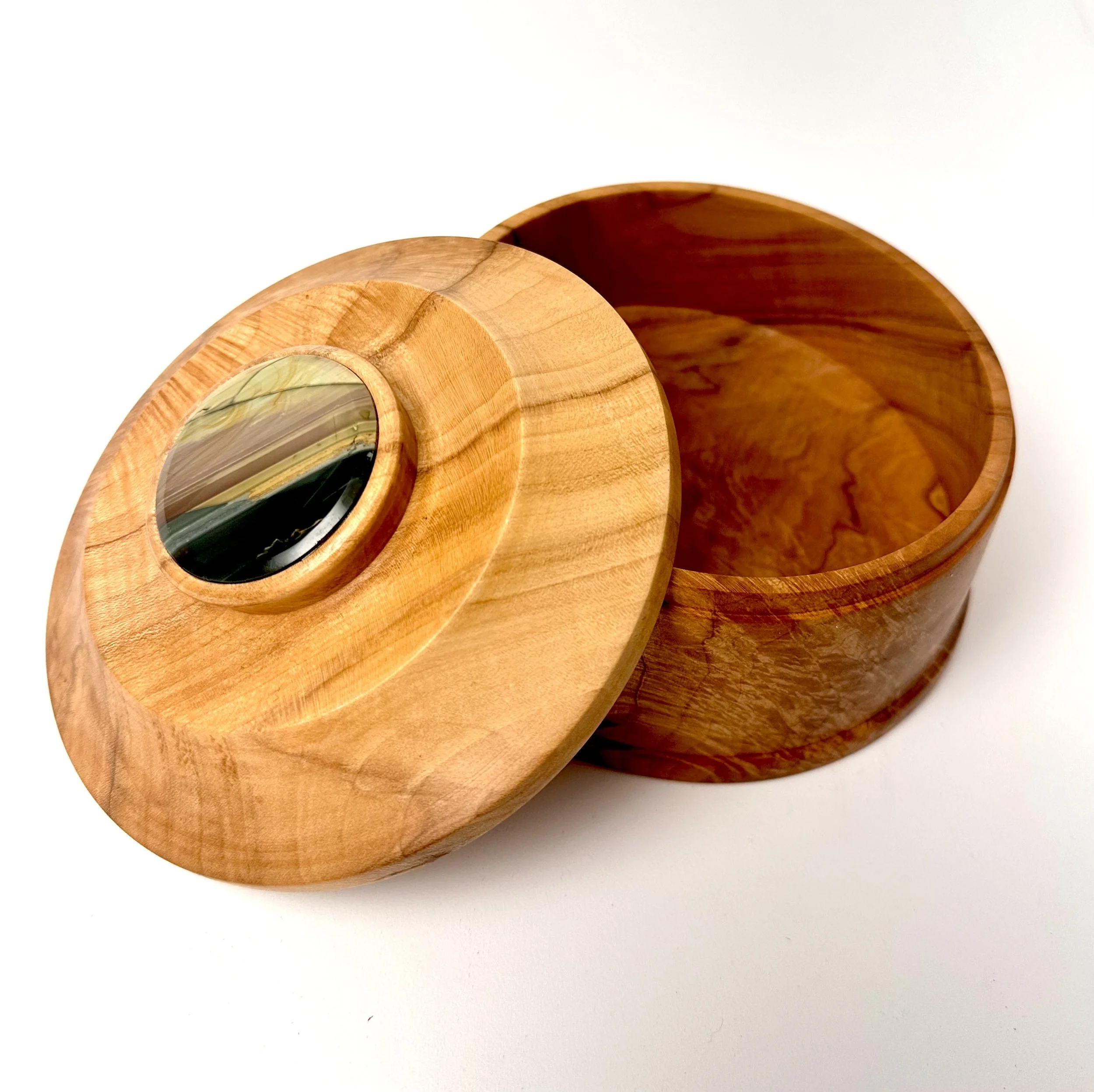 Round wooden box with a removable lid, featuring a black decorative element in the center of the lid, made of smooth wood with visible grain patterns.