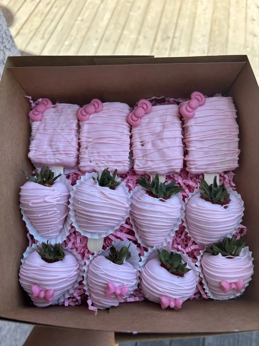 A box of assorted strawberries dipped in pink chocolate, decorated with pink bows and beads, arranged on pink shredded paper.