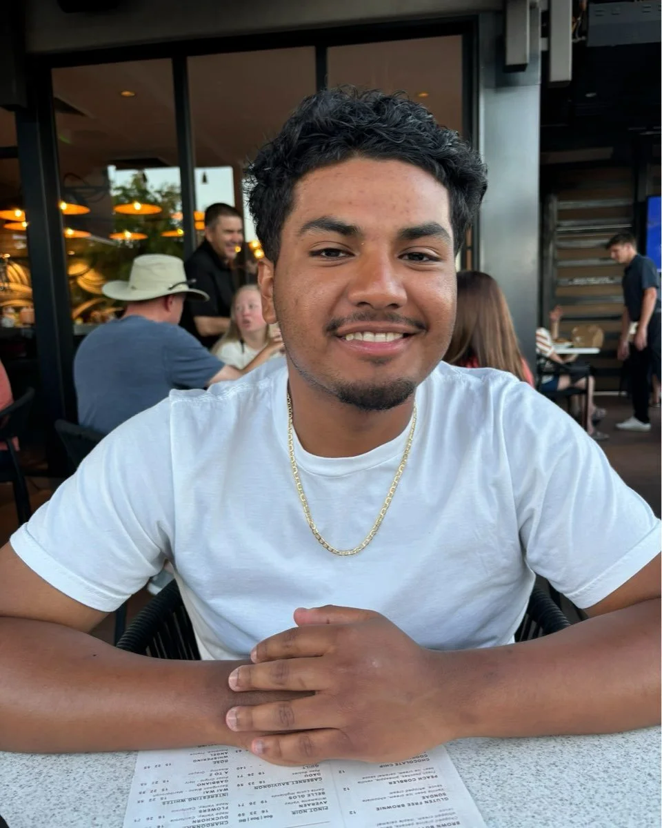 Young man with curly black hair and a goatee, smiling at the camera, wearing a white T-shirt and gold chain, sitting at a restaurant table with a menu in front, in a lively indoor setting with other diners and a modern decor.