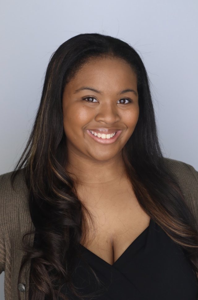 Young woman with long, dark hair and a warm smile, wearing a black top and brown cardigan, posing against a plain light gray background.