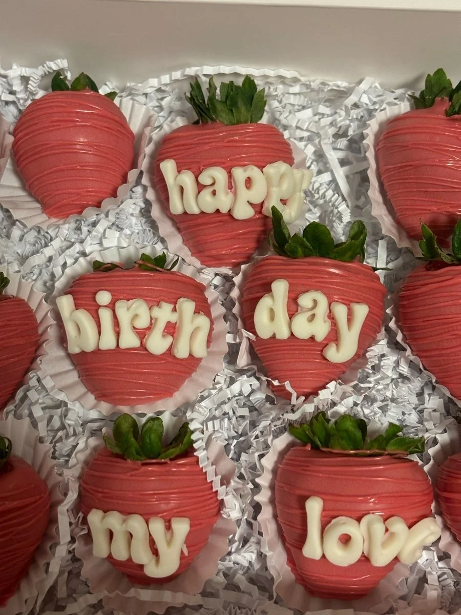 Strawberries dipped in pink chocolate with white chocolate writing saying 'happy birthday love' on some of them, arranged on a bed of white crinkled paper