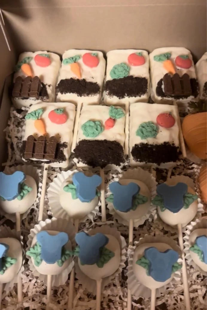 Assorted decorated cake pops in a box, featuring designs with carrots, green leaves, red circles, and brown chocolate pieces.
