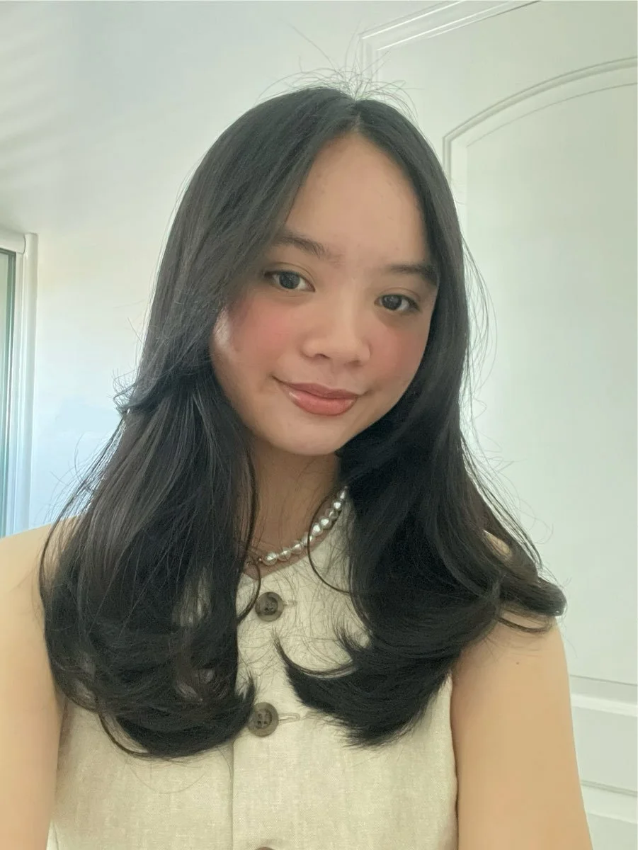 A young woman with long dark hair, smiling, wearing a cream-colored sleeveless top with large buttons and a pearl necklace, standing in front of a white door and wall.