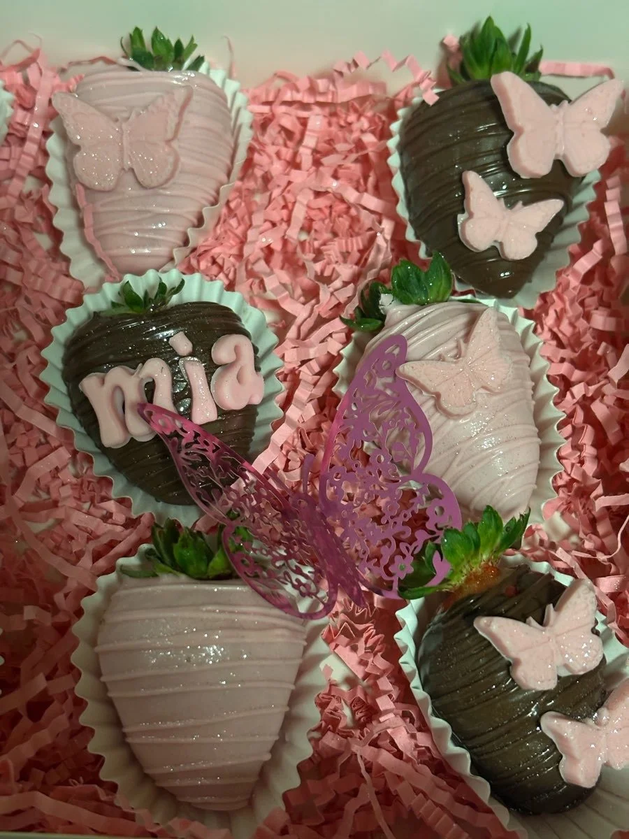 Decorated strawberries with pink and dark chocolate coatings, adorned with pink butterfly-shaped decorations and small pink fondant butterflies, placed on pink shredded paper.