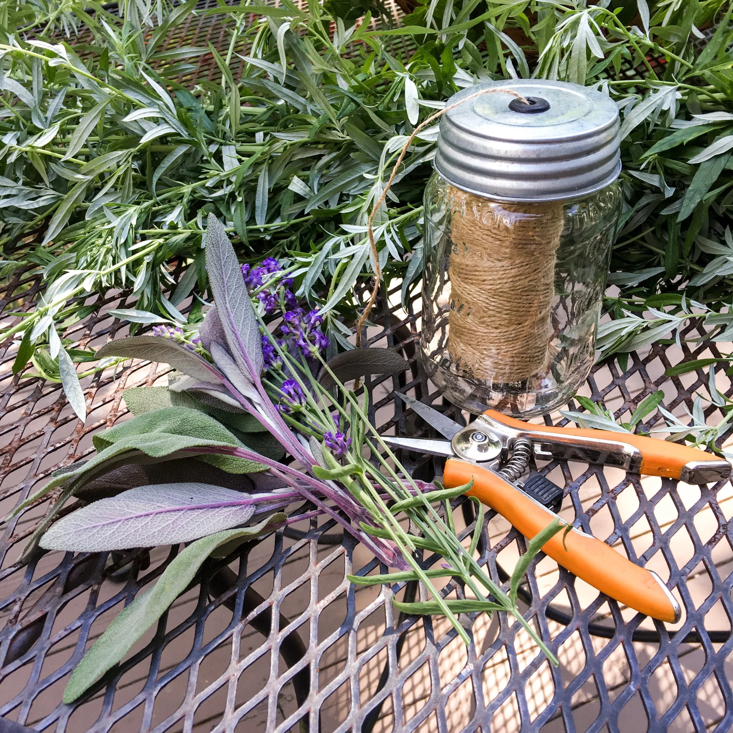 A metal outdoor table with gardening supplies including a bunch of lavender and sage, a glass jar with twine on a spool inside, and orange-handled pruning shears, with green shrubbery in the background.