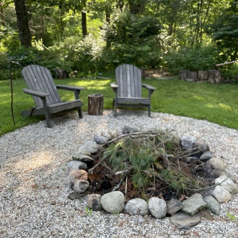 Two gray wooden Adirondack chairs facing inward around a fire pit made of rocks, with a small tree stump between them, in a backyard with green grass and trees.