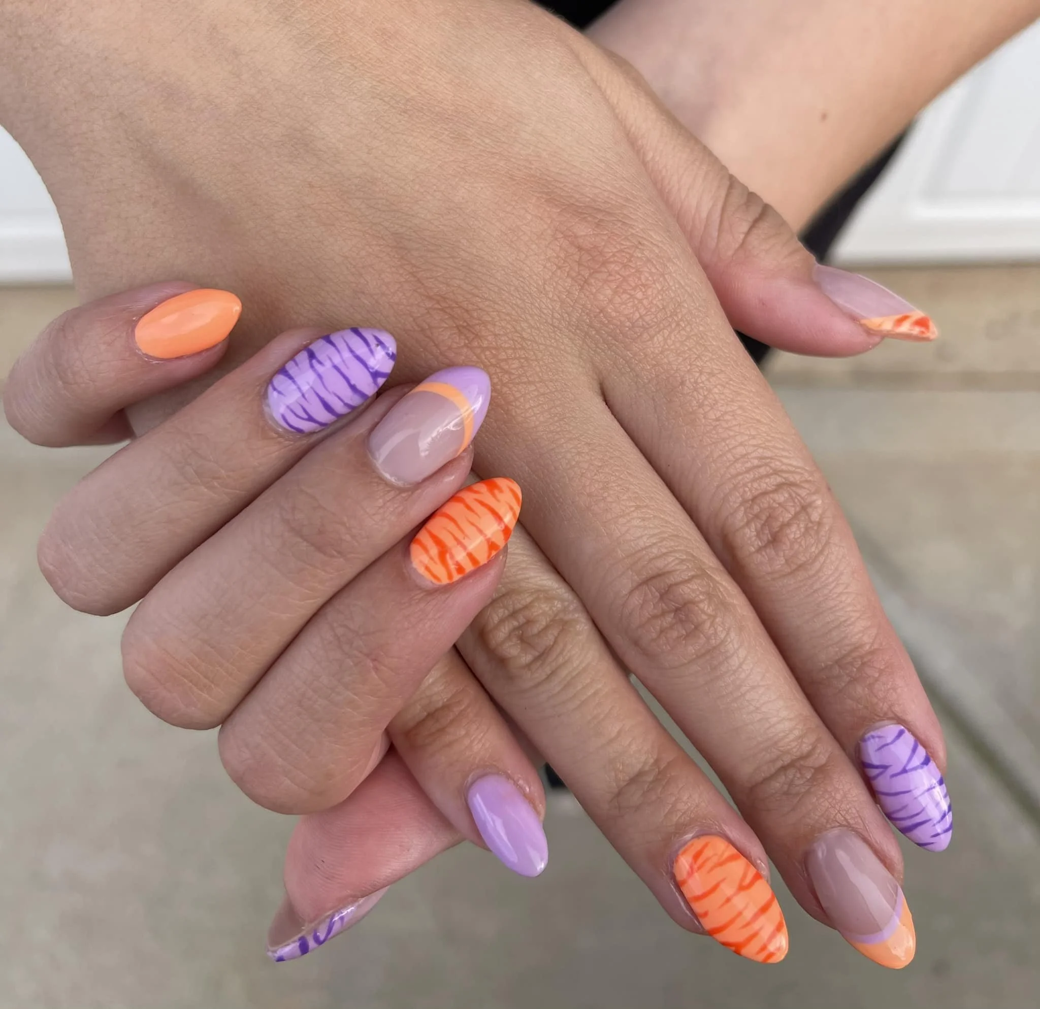 Close-up of hands with brightly colored and patterned nail polish, featuring purple, orange, and clear nails with different designs.