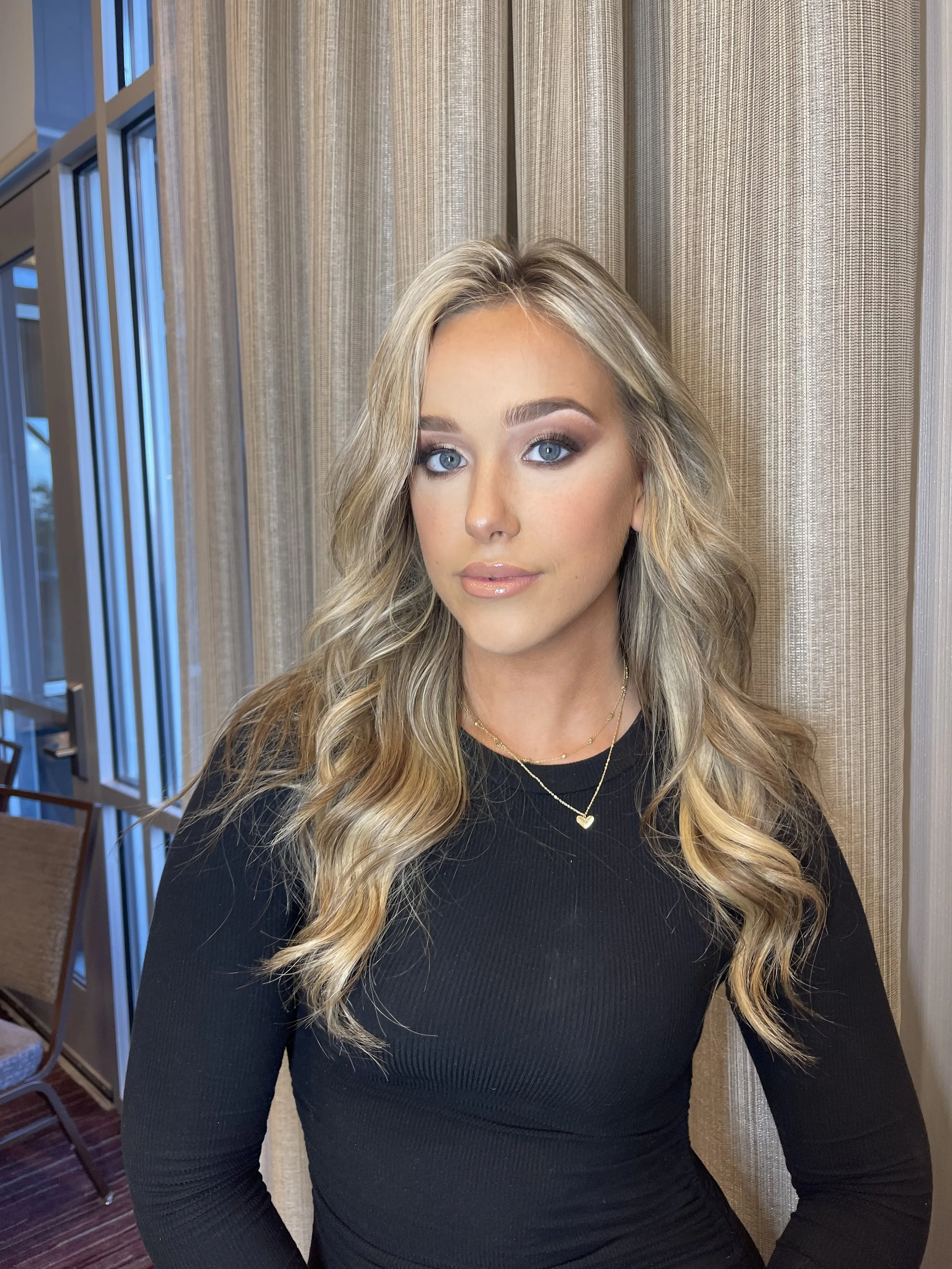 A young woman with long, wavy blonde hair and blue eyes, wearing a black long-sleeve top and gold jewelry, posing indoors in front of beige curtains.