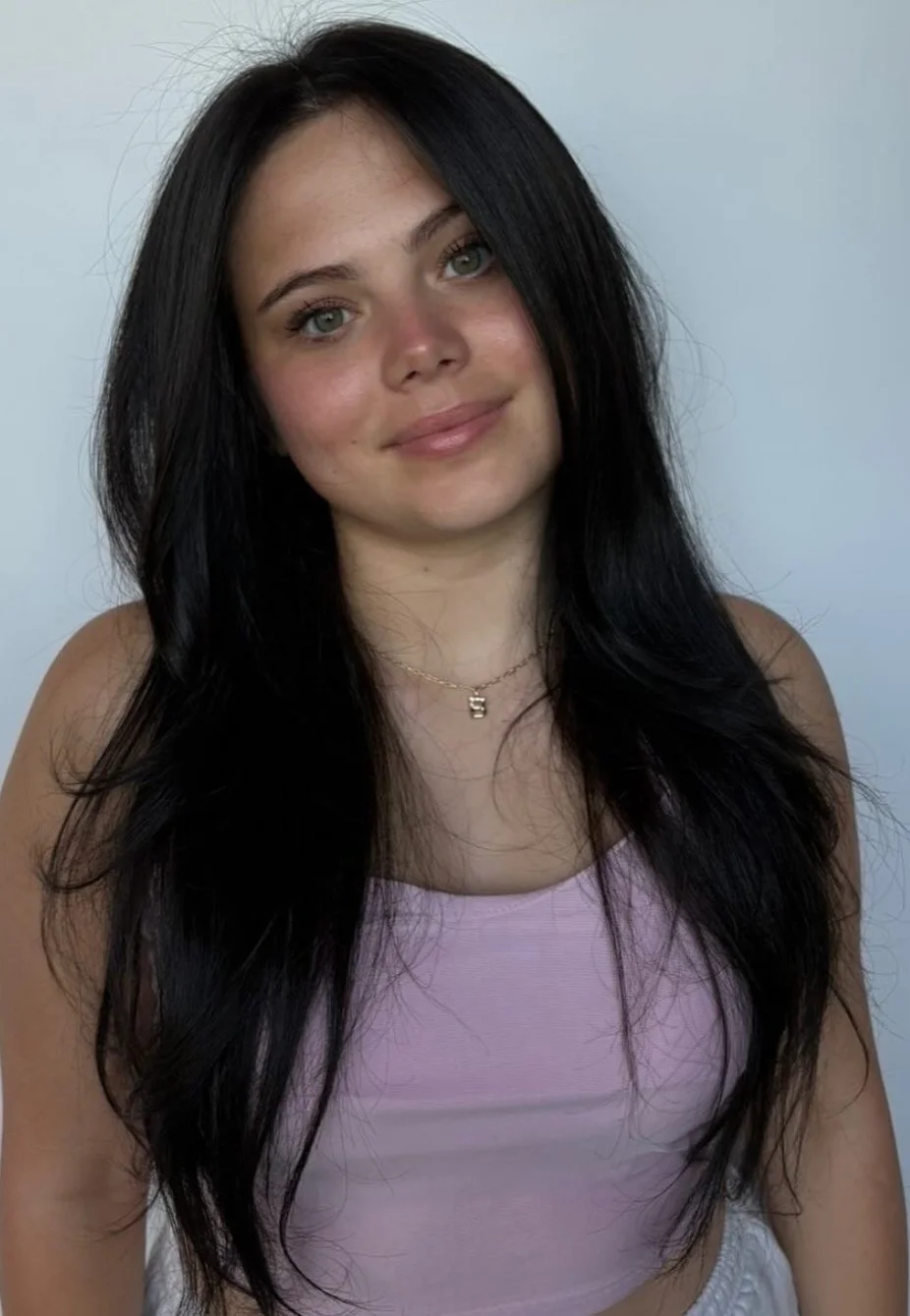 A young woman with long black hair, light skin, and blue eyes, smiling softly, wearing a pink sleeveless top and a delicate chain necklace with a small pendant, posing against a plain light background.