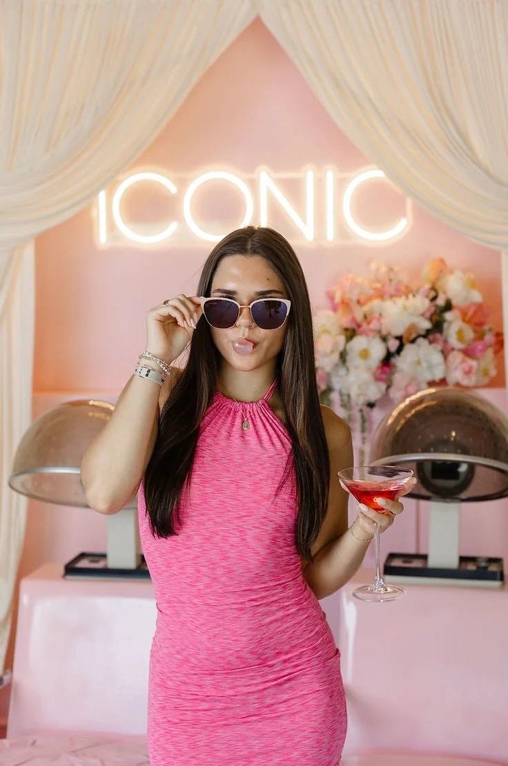 Young woman with long brown hair wearing a pink dress and sunglasses, holding a cocktail glass, standing in a pink decorated room with a neon sign that says 'ICONIC' in the background.