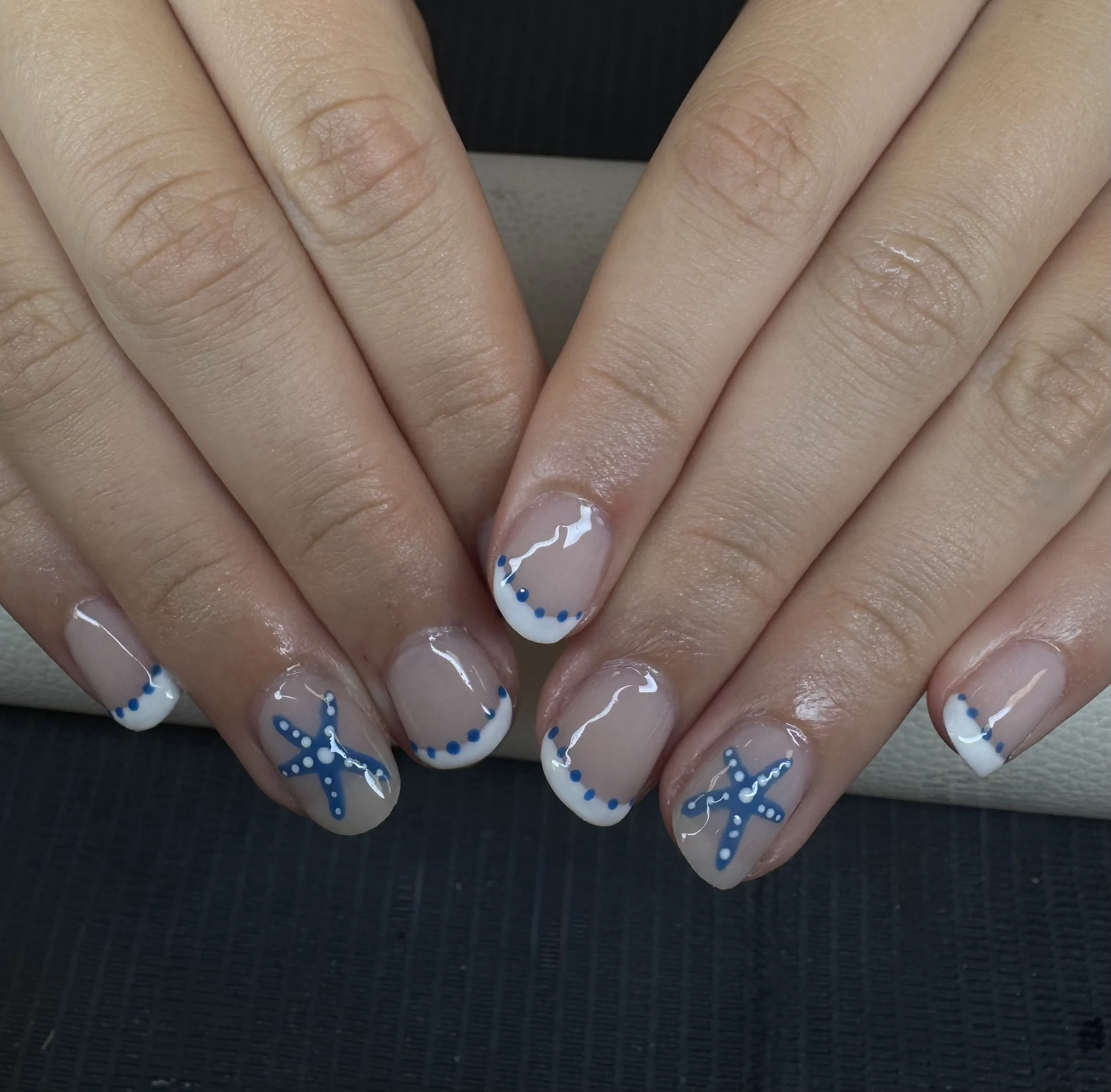 Hands with manicured nails featuring nautical-themed nail art, including white tips decorated with blue dotted borders and blue starfish and an anchor design.