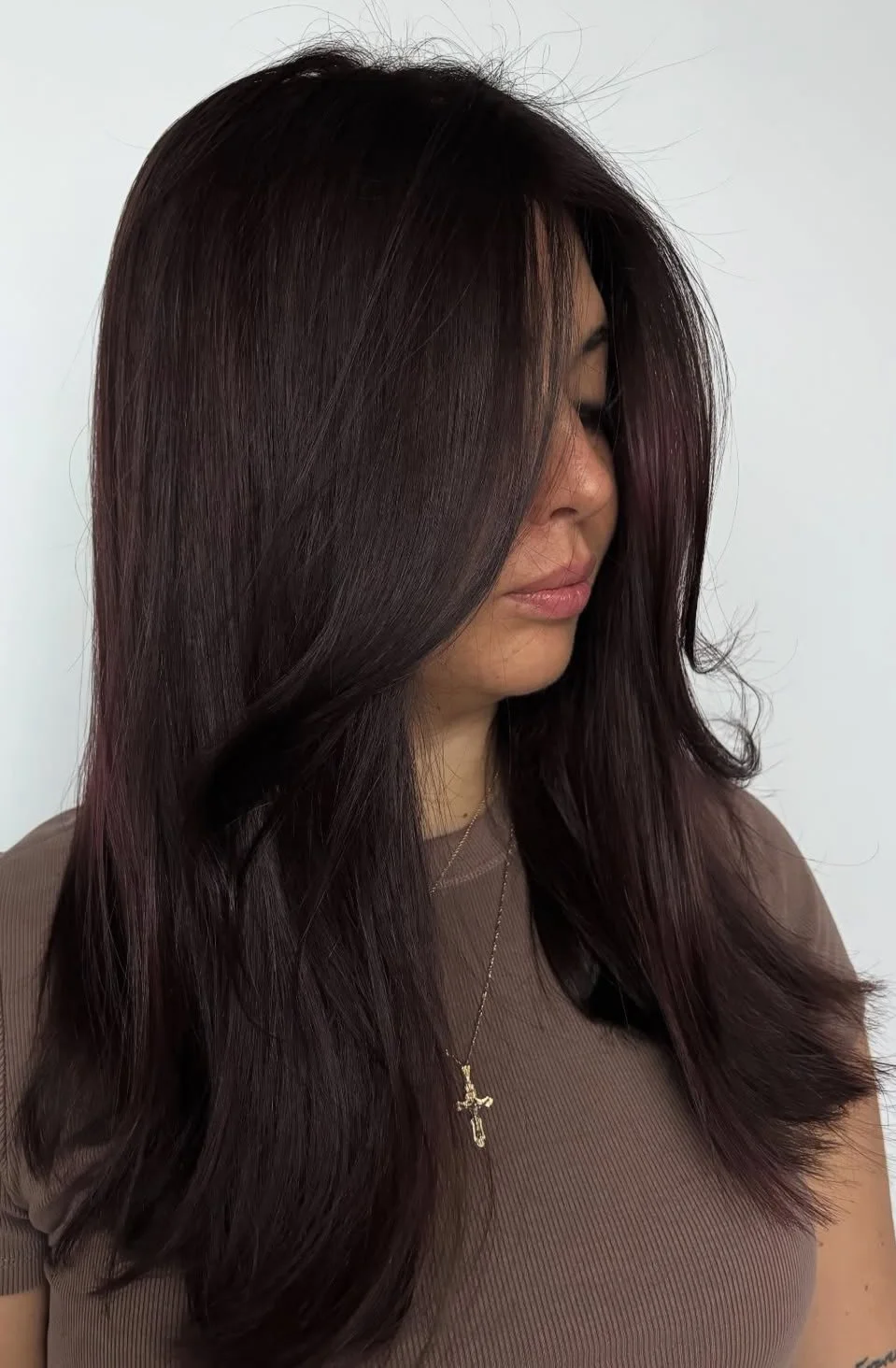 A woman with long, dark, and shiny hair, wearing a brown top and a gold cross necklace, with her eyes closed and head tilted downward against a plain white background.