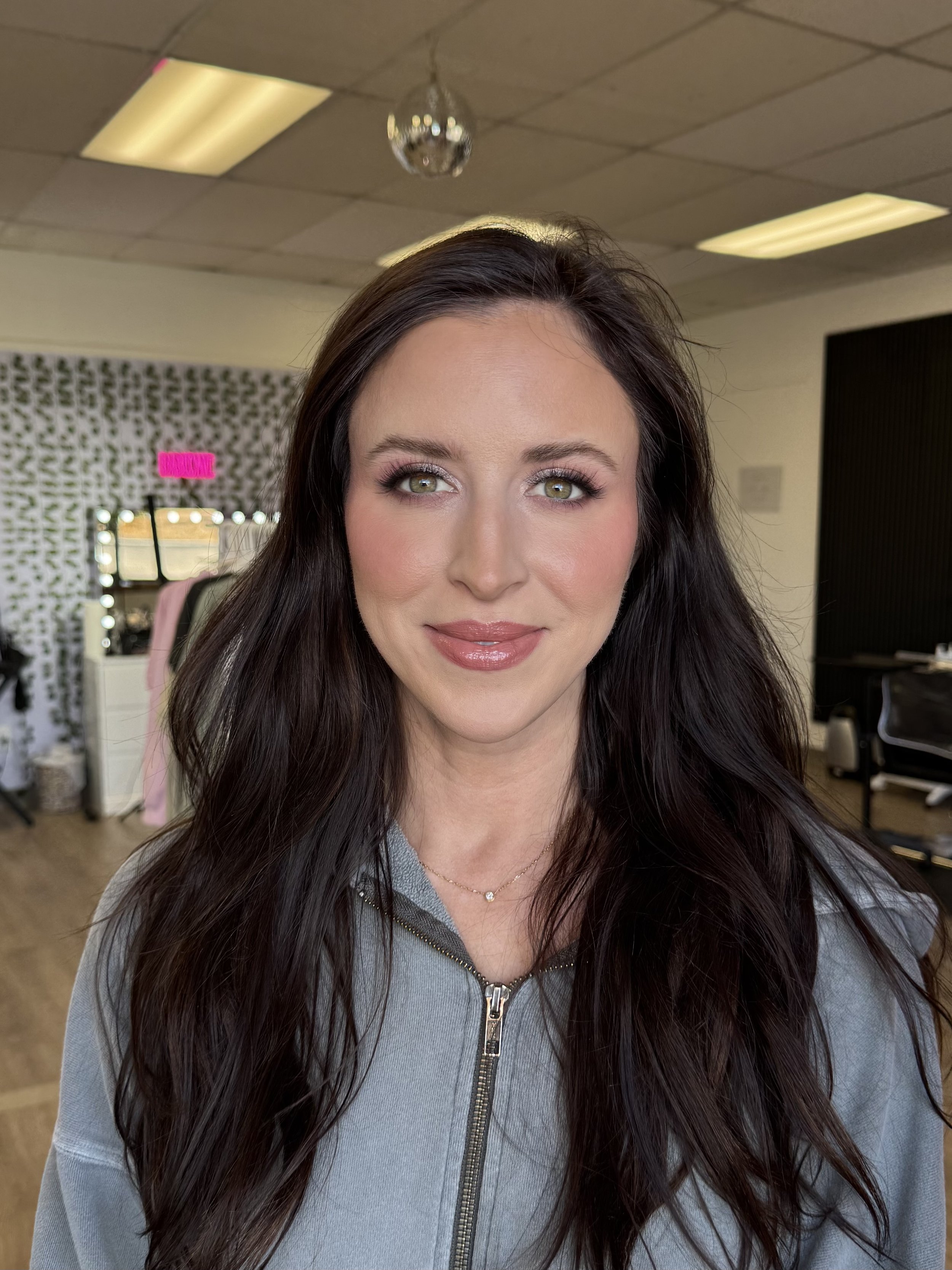 A woman with long dark hair and makeup, smiling, wearing a gray zip-up top and a delicate necklace, in a studio or salon setting.