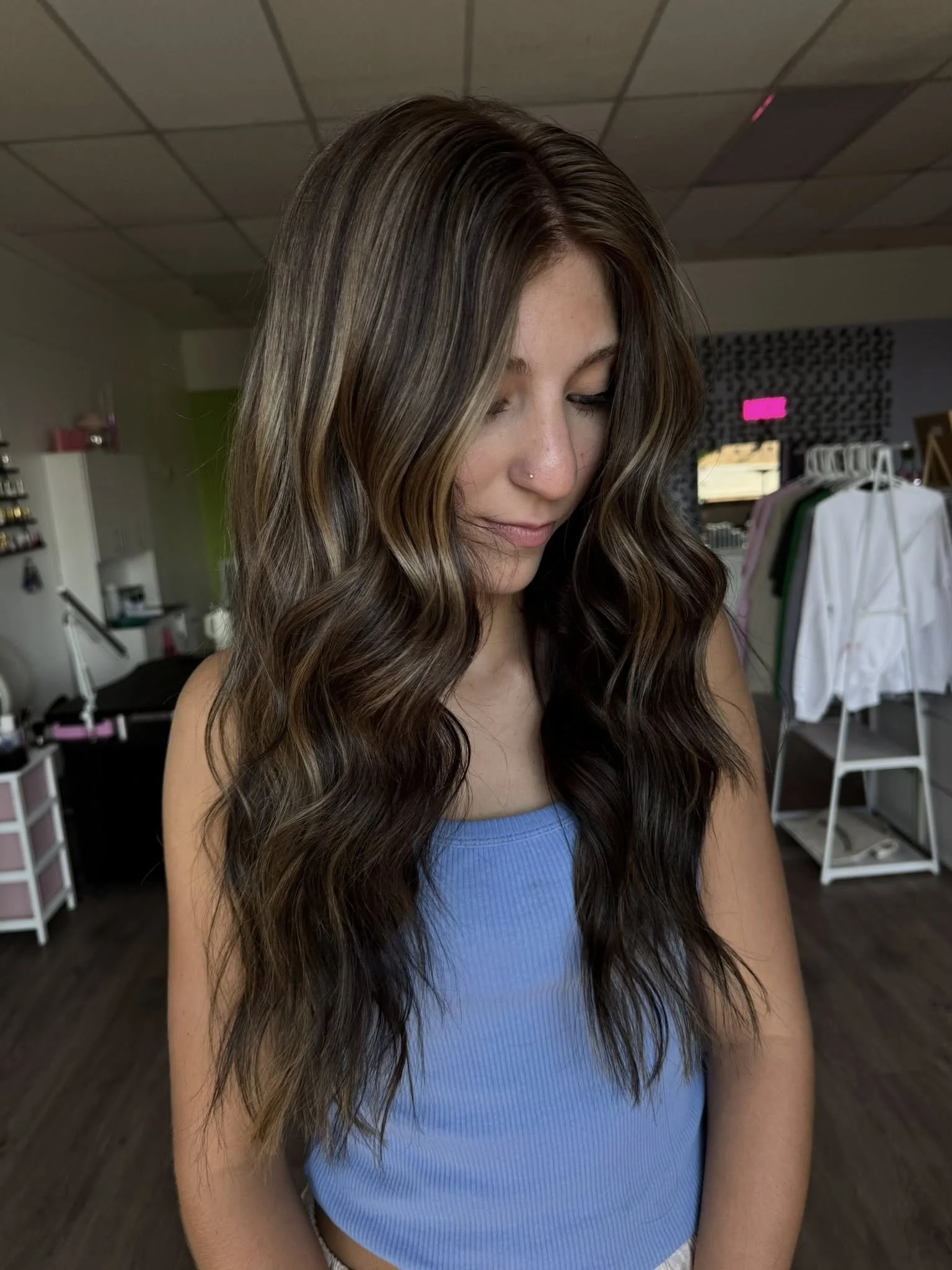 A young woman with long, wavy brown hair with blonde highlights, wearing a light blue crop top, standing in a salon or boutique.