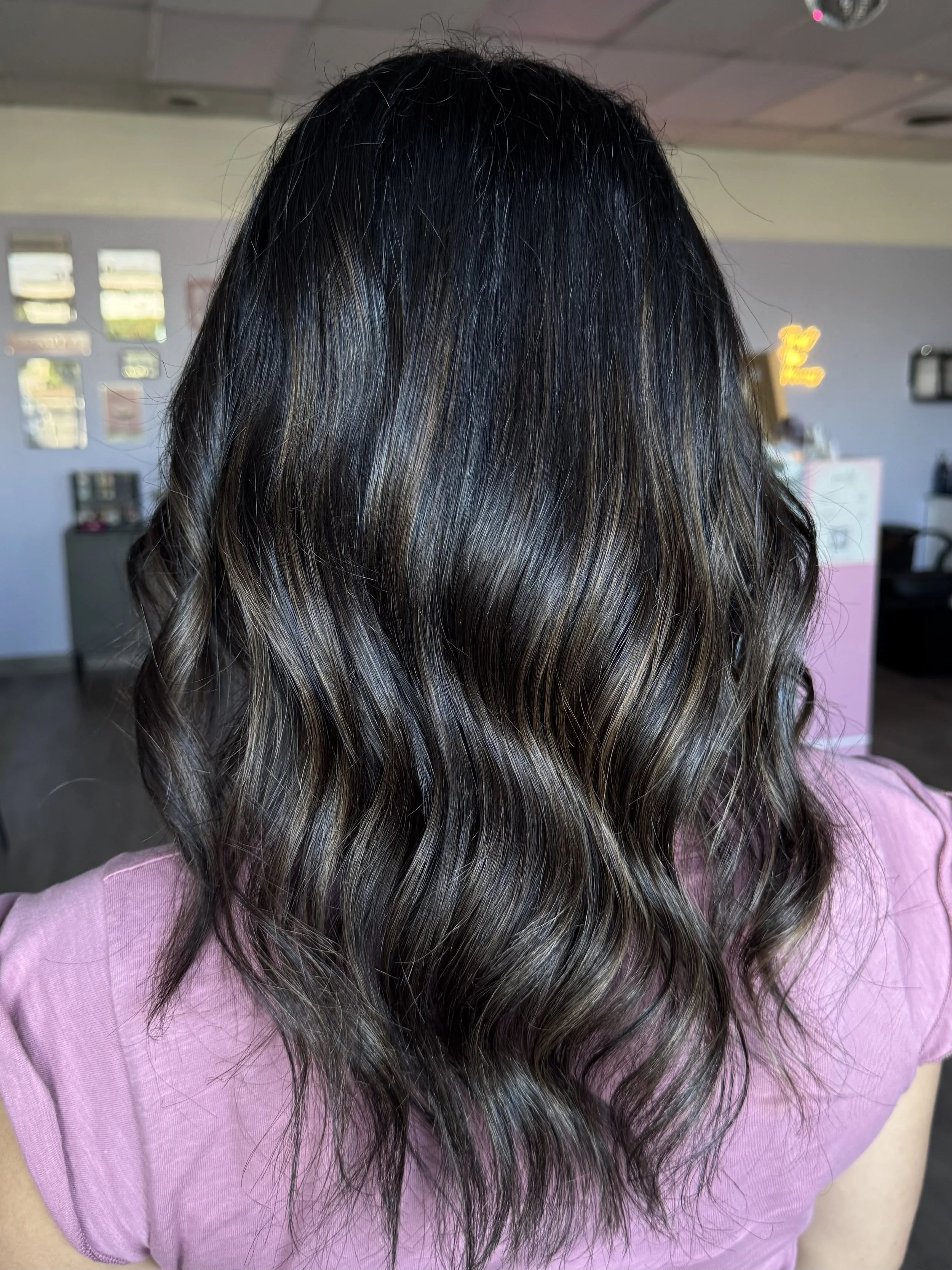 Back of a person's head showing dark brown wavy hair with subtle highlights, wearing a pink shirt in a salon or studio setting.
