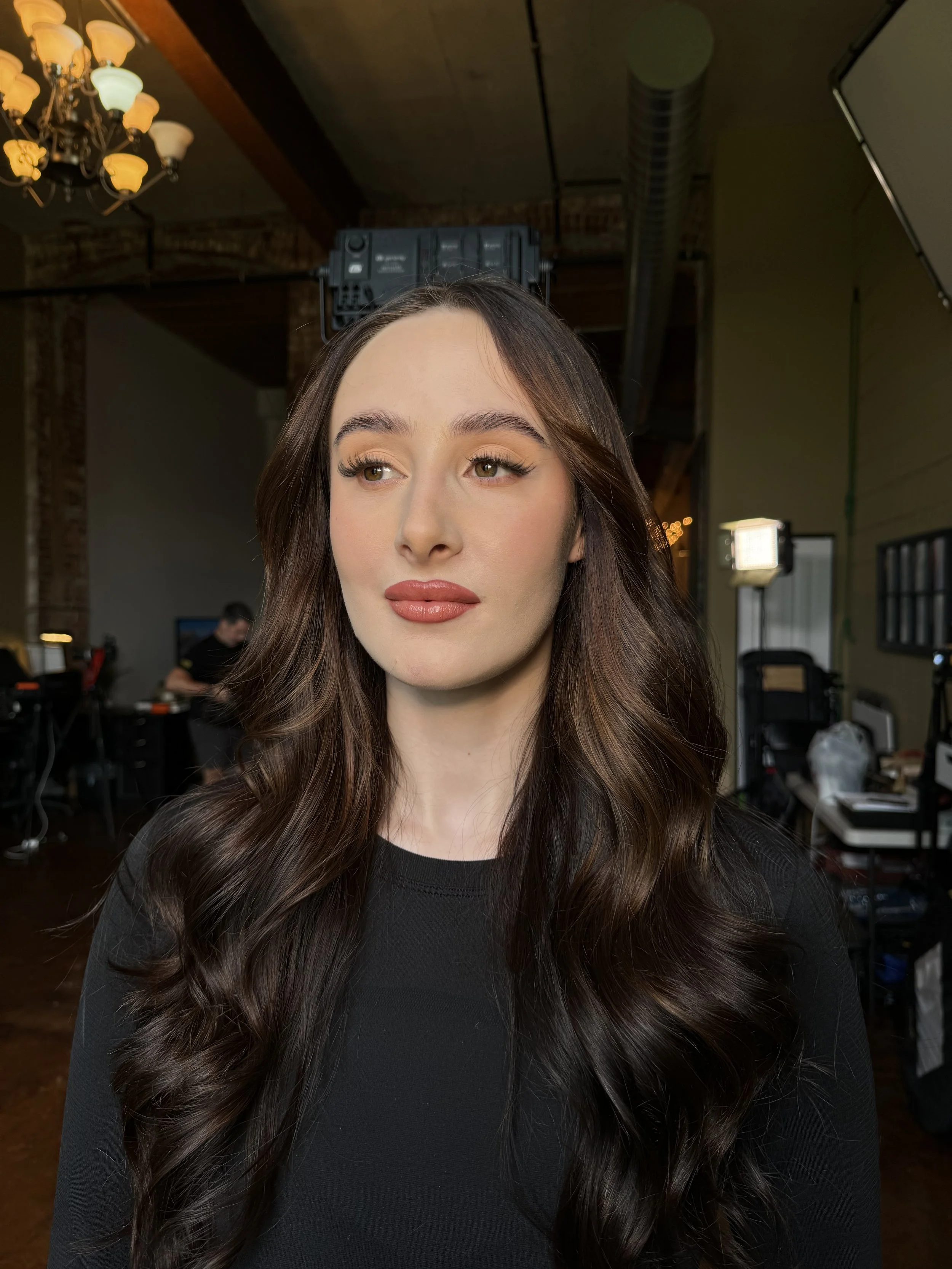A woman with long, wavy brown hair and makeup standing in a photography studio with lighting equipment and a photographer in the background.