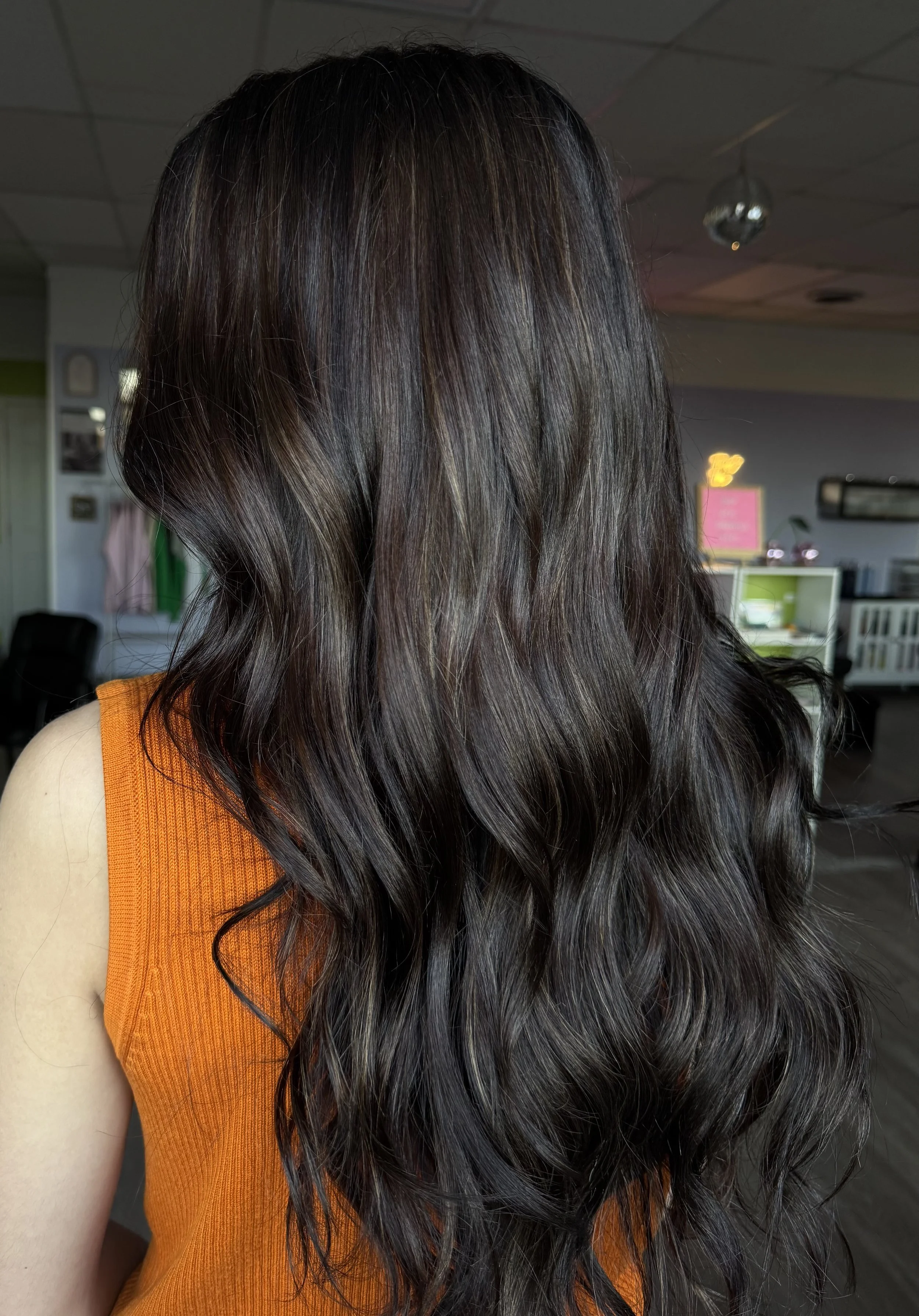The image shows a woman with long, wavy, dark brown hair wearing an orange sleeveless top, standing in an indoor space with a ceiling decorated with a small disco ball and various objects in the background.
