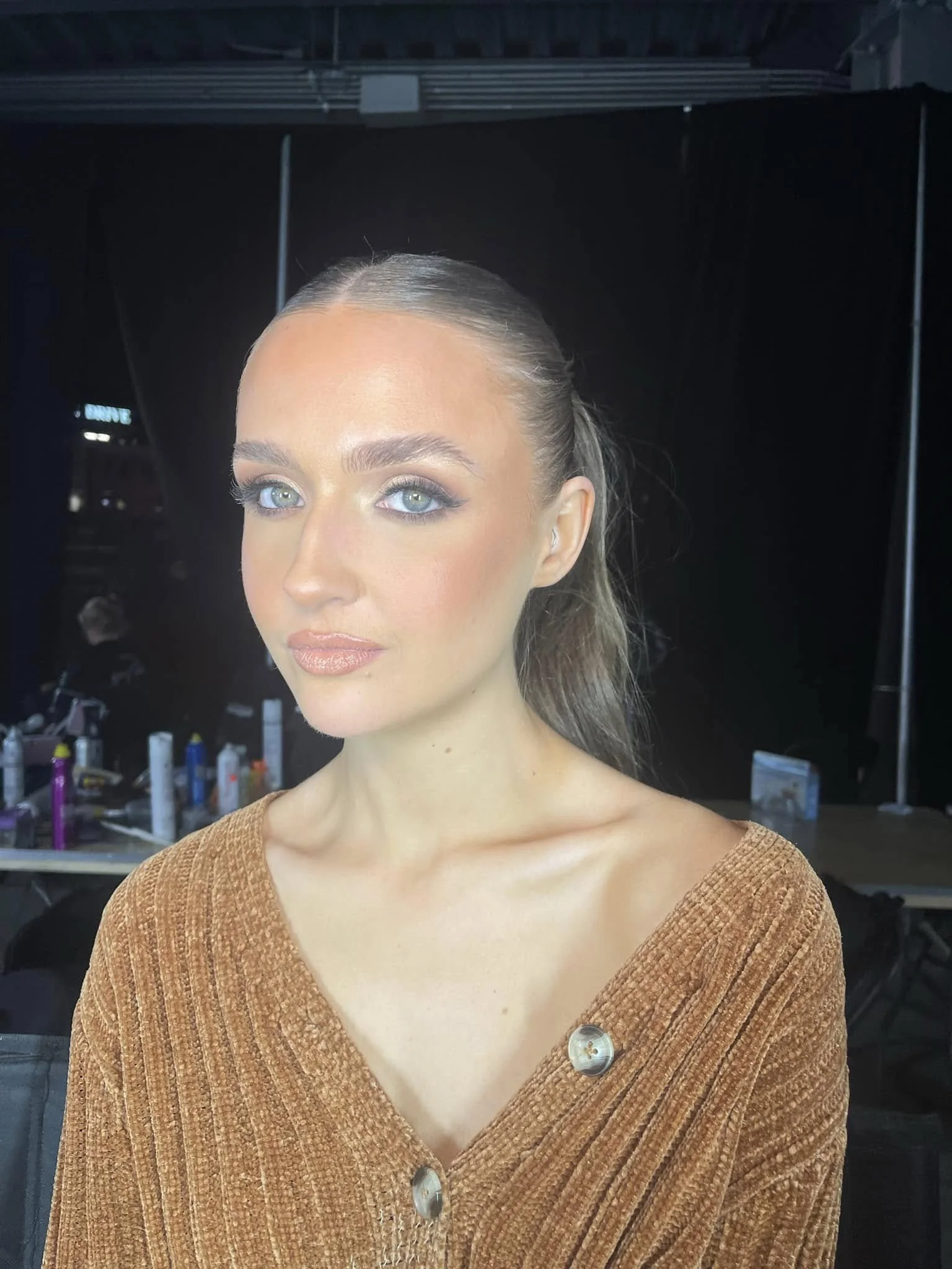 A woman with long brown hair styled in a ponytail, blue eyes, and makeup, wearing a brown textured button-down shirt, standing in a studio setting.