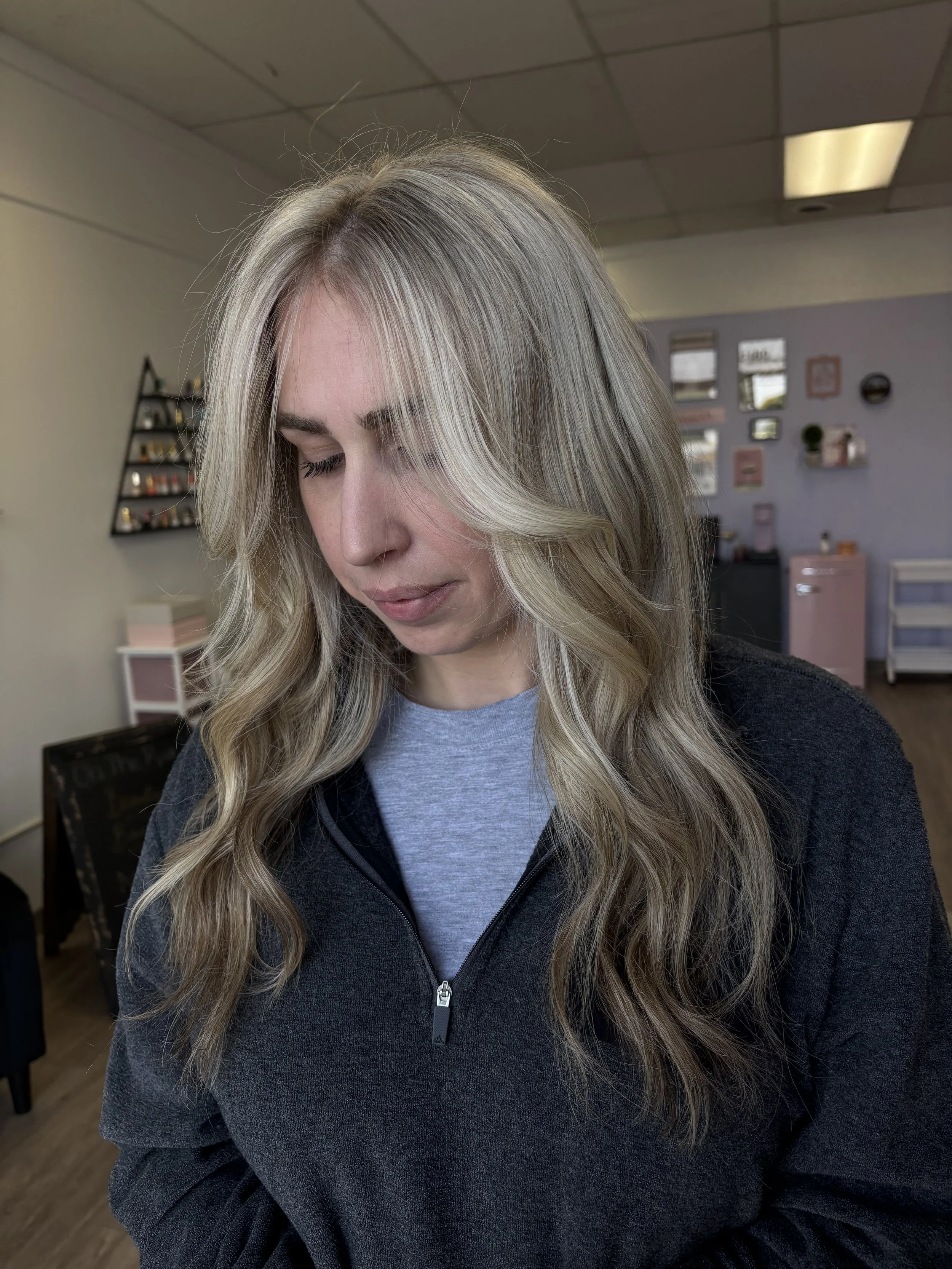 A woman with long, wavy blonde hair and light skin is looking down in a room with pastel-colored decor and small framed pictures on the wall.