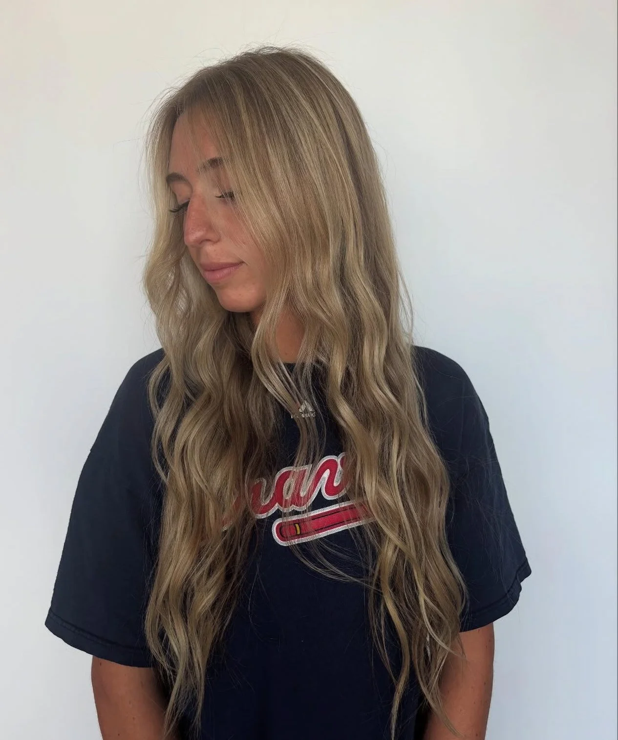 A woman with long wavy blonde hair and light skin wearing a black T-shirt with red and white lettering standing against a plain white wall.