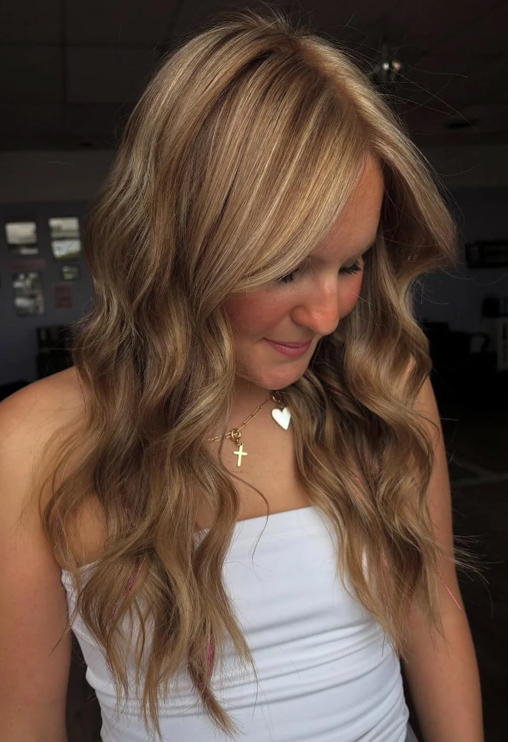 A woman with long, wavy blonde hair looking downward, wearing a white top and layered necklaces with a cross and heart pendant, in an indoor setting.