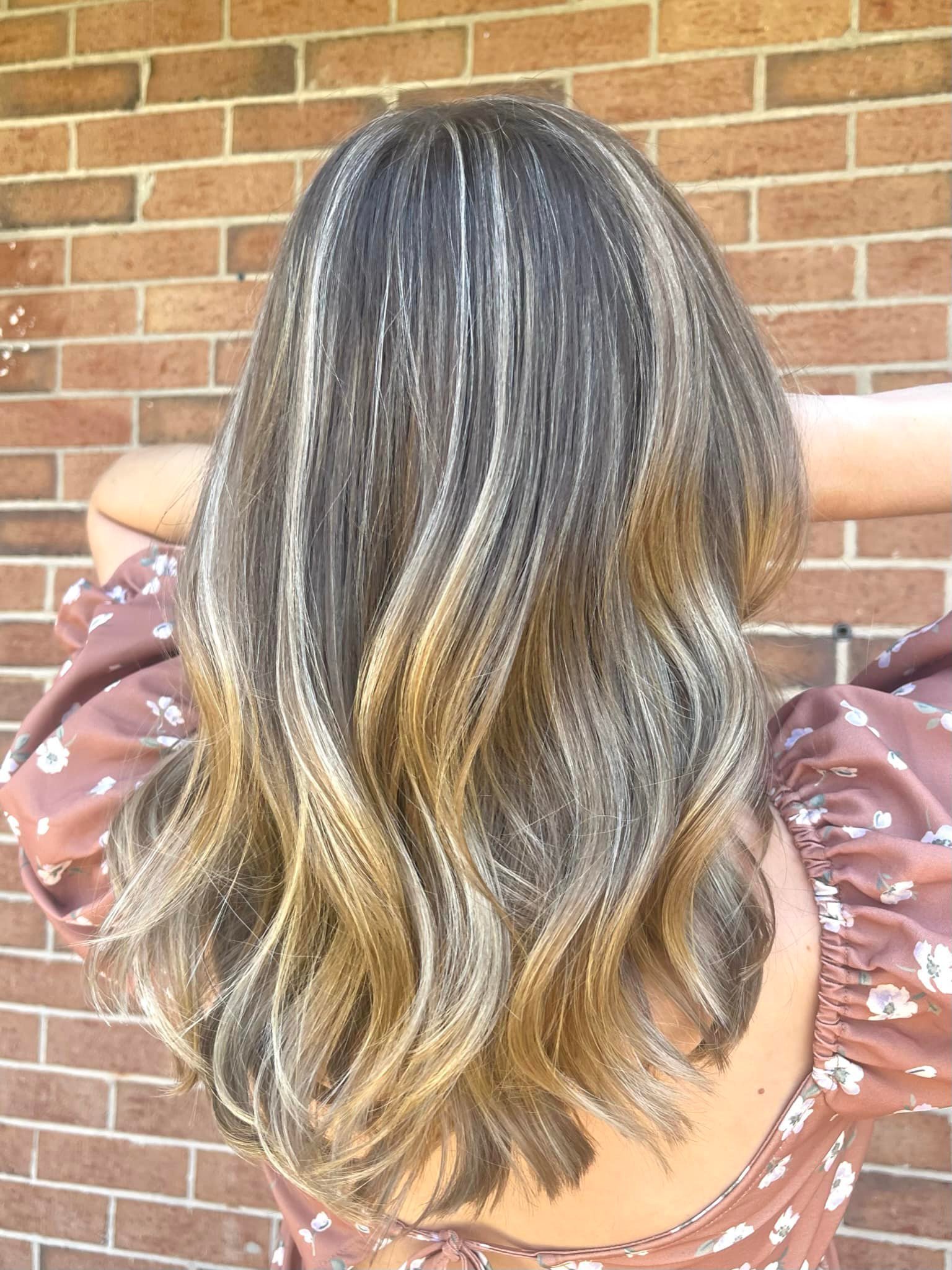Back view of a woman's shoulder-length, wavy blonde hair with darker roots, standing in front of a brick wall. She is wearing a pink floral dress with puffed sleeves.