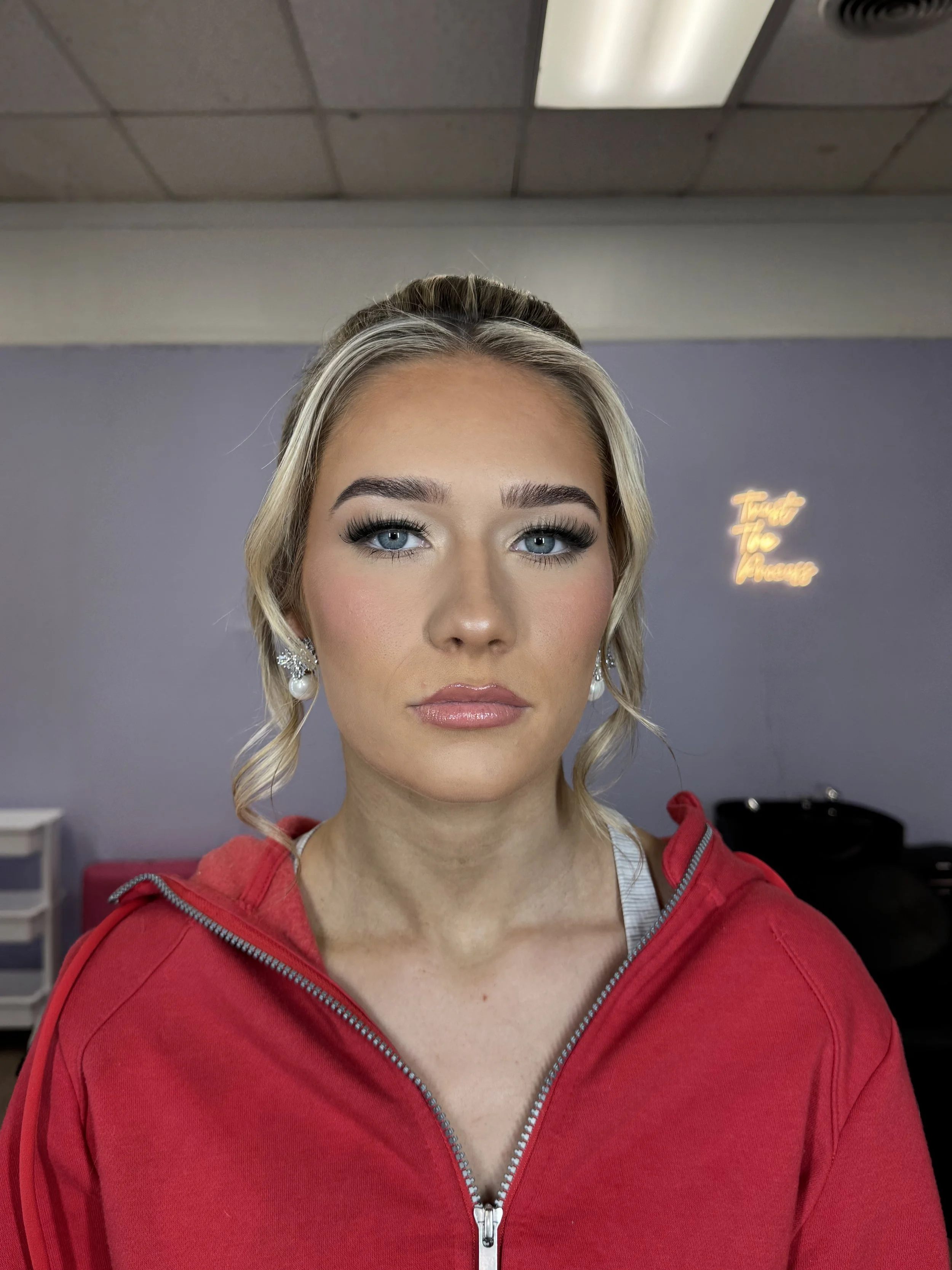 A woman with blonde hair, blue eyes, and makeup, wearing a red zip-up hoodie, looking directly at the camera, in a room with a purple wall and a neon sign that says 'Trust The Process' in the background.