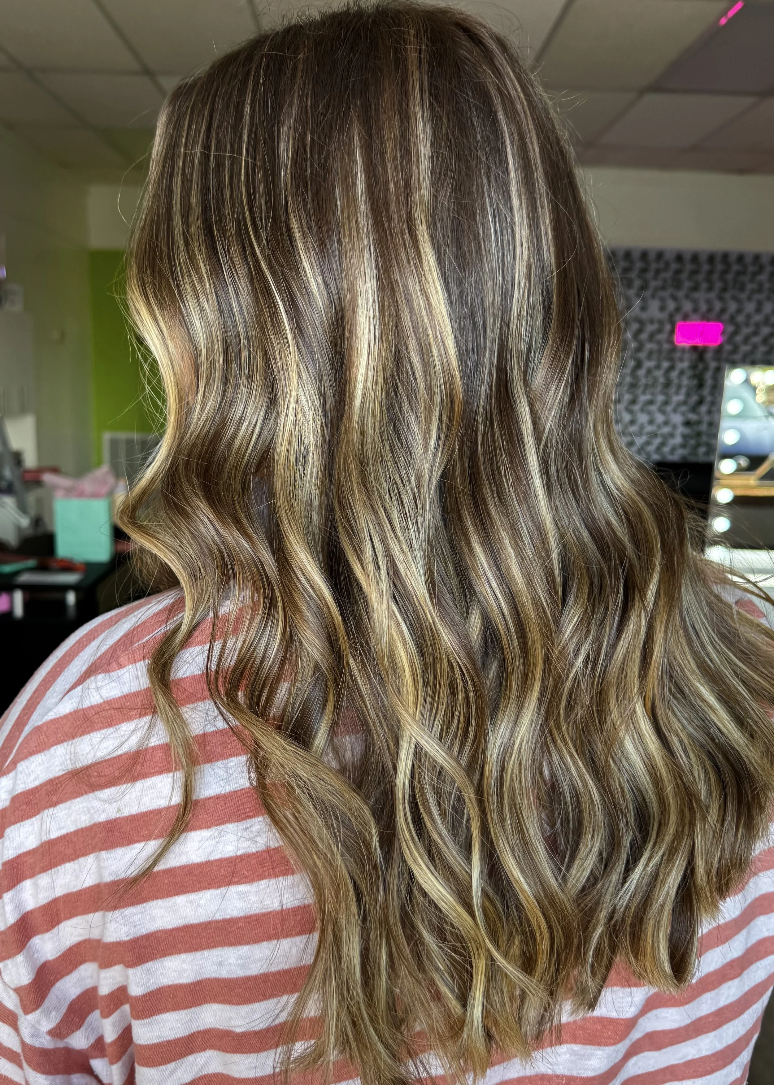 A person with long, wavy, blonde hair featuring highlights and lowlights. The person is wearing a pink and white striped shirt and is in an indoor setting, possibly a hair salon.