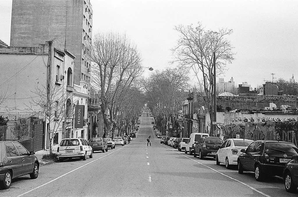 Rua urbana com carros estacionados de ambos os lados, árvores sem folhas e prédios ao fundo, em um dia nublado.