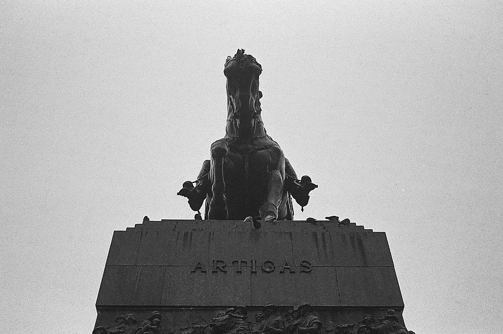 Estátua de um cavalo com soldados, com o nome "Artigas" na base, vista de baixo para cima em uma fotografia em preto e branco.
