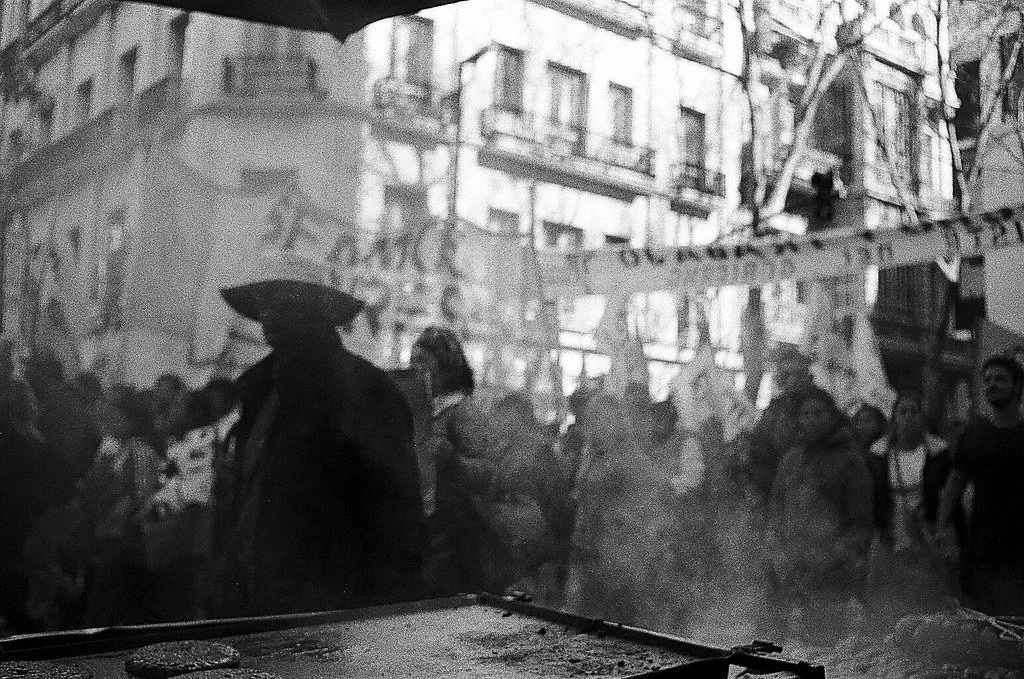 Pessoa com guarda-chuva caminhando em uma rua movimentada com vários pedestres, edifícios ao fundo, em clima chuvoso.