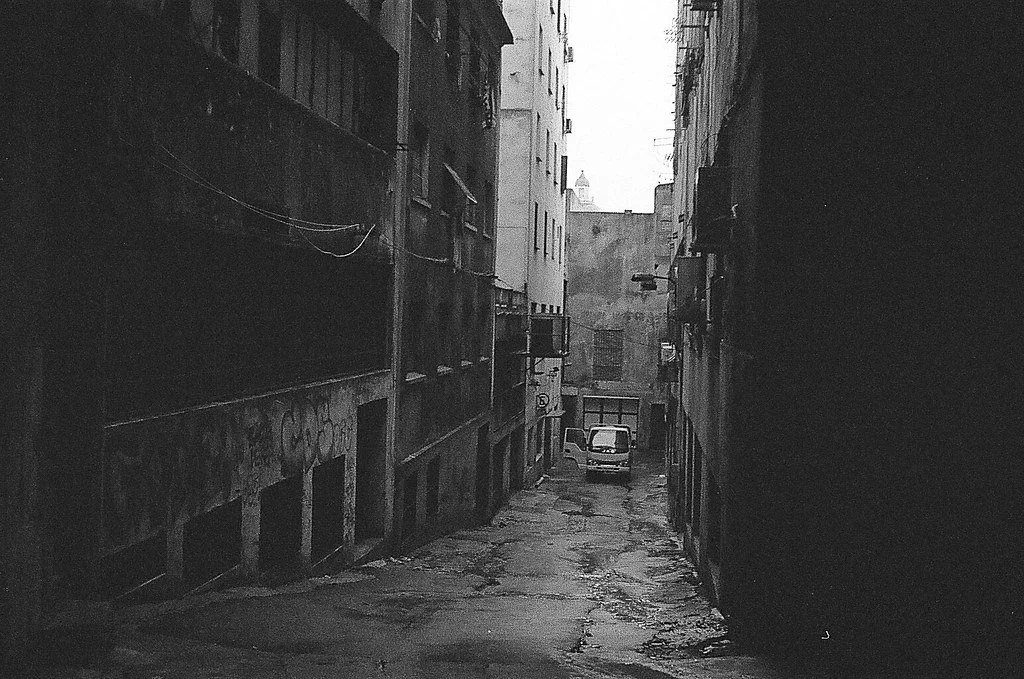 Rua estreita entre prédios antigos, com carro estacionado ao fundo, em um ambiente urbano sombrio.