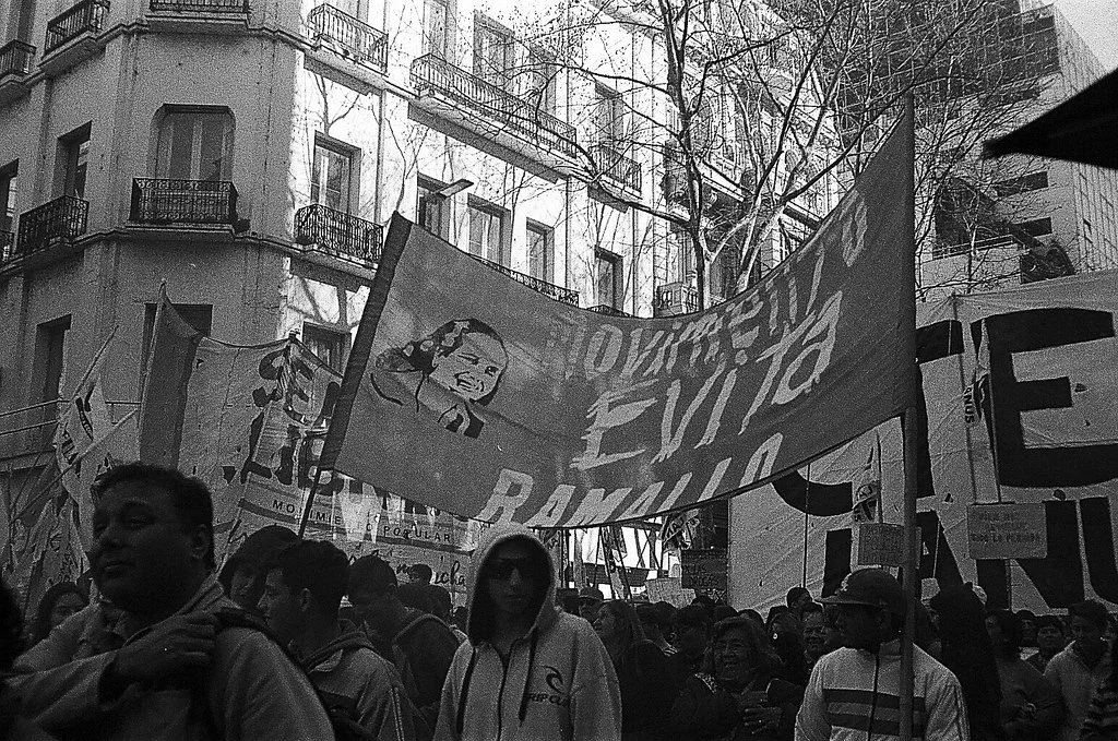 Protesto na rua com várias pessoas, banners e faixas, incluindo uma com uma caricatura de uma mulher e com texto "EVIÇÃO" e "RACISMO" em uma rua urbana com prédio ao fundo.