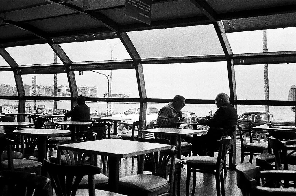 Interior de um restaurante ou café com várias mesas e cadeiras, duas pessoas sentadas numa mesa conversando, uma pessoa olhando para fora pela janela, no ambiente de dia.