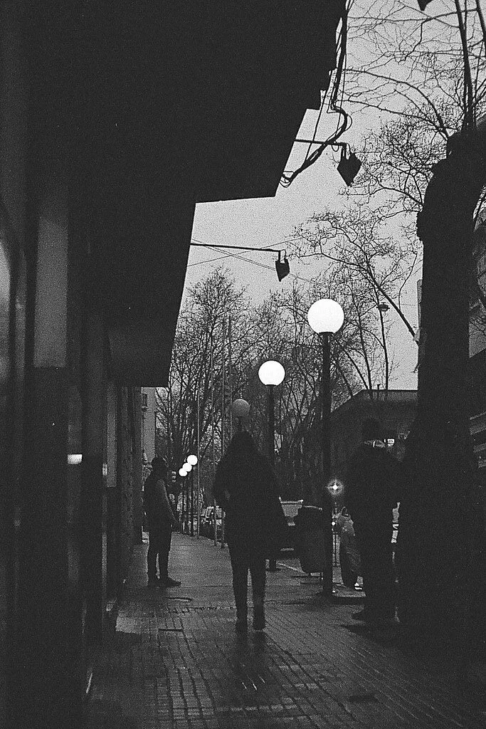 Rua urbana à noite com pessoas caminhando, árvores sem folhas e postes de iluminação com globos de luz, em uma cena de outono ou inverno.