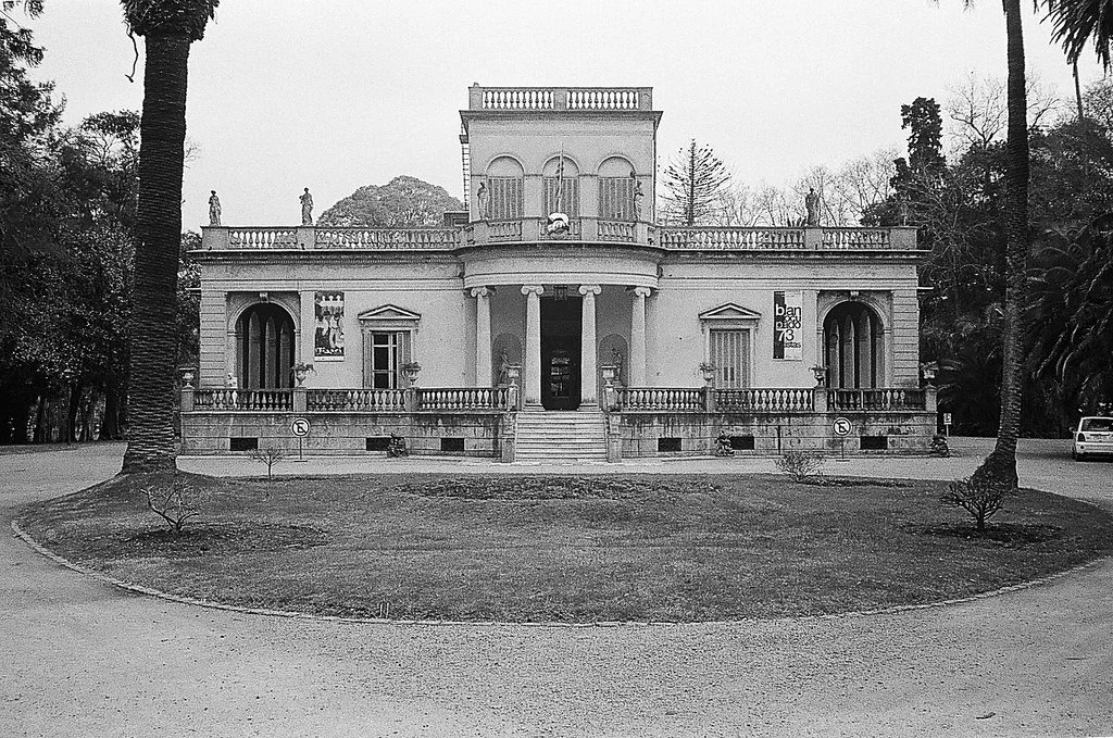 Uma mansão histórica de estilo clássico com balcões, colunas e estátuas no telhado, cercada por árvores e um jardim, vista em preto e branco.