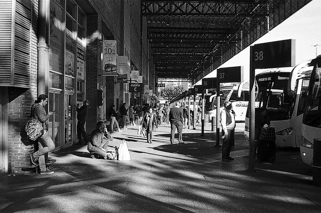 Cena de uma estação de ônibus com pessoas esperando, algumas sentadas e outras de pé, e ônibus estacionados em plataformas.