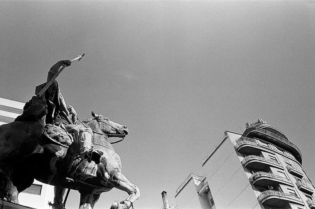 Estátua de um cavaleiro em uma praça urbana com edifícios modernos ao fundo, em preto e branco.