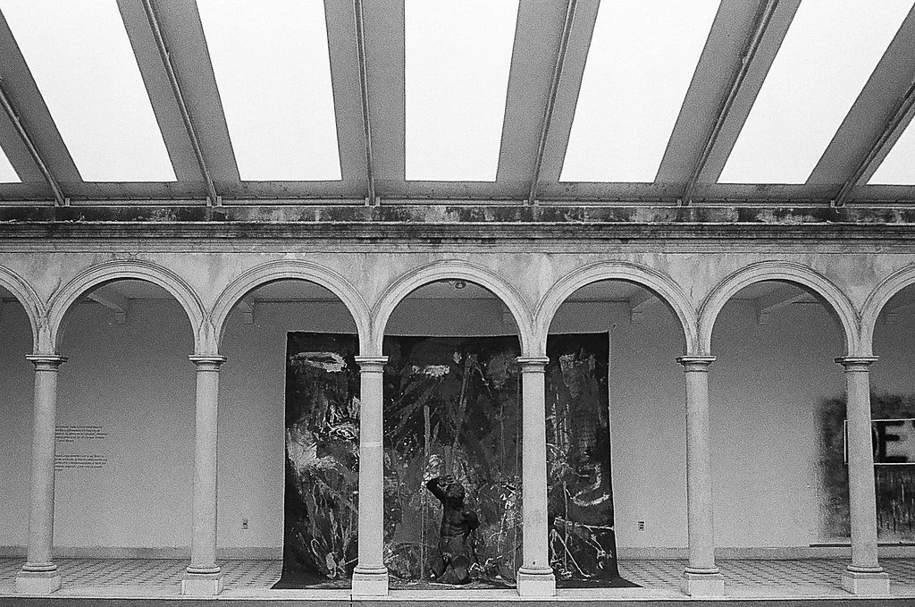 Escultura de uma pessoa em meio a uma instalação artística com painéis de fundo, localizada em uma galeria de arte com arcos e teto de vidro.