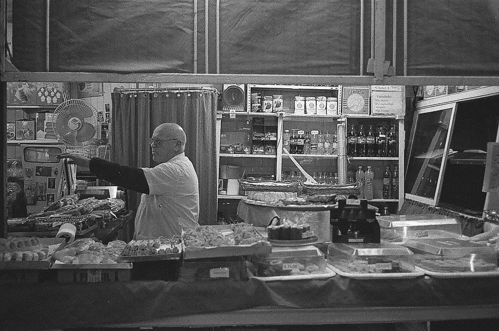 Homem vendendo doces e bebidas em uma mercearia ou boteco tradicional, com prateleiras cheias de produtos ao fundo.