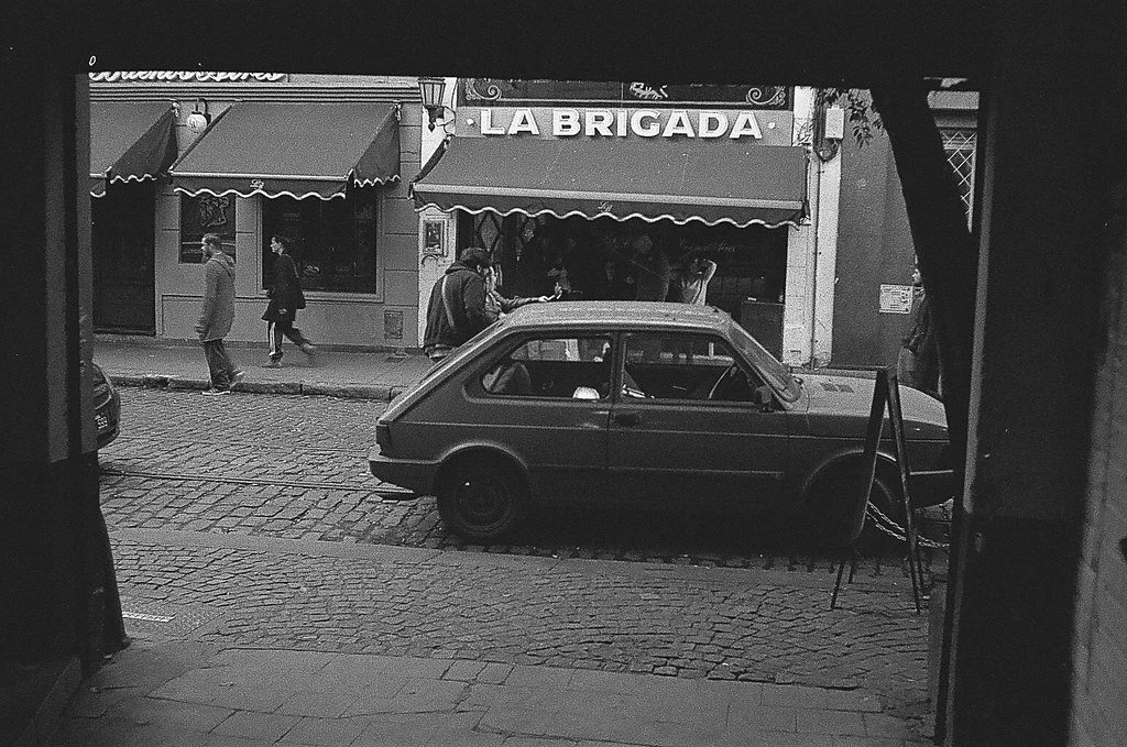 Cena de rua à noite vista de uma janela, com carro estacionado na calçada, pessoas caminhando e uma loja chamada 'La Brigada' ao fundo.