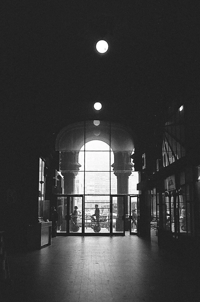 Imagem em preto e branco do interior de uma estação de metrô com vista para o exterior, através de janelas grandes com arcos. Pessoas e motocicletas podem ser vistas do lado de fora, enquanto dentro há silhuetas de pessoas e estruturas arquitetônicas