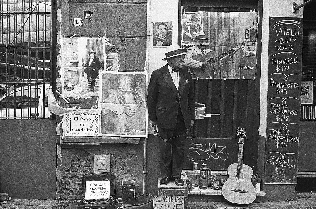 Homem vestido de terno preto, óculos e chapéu, tocando violão na rua em frente a uma fachada com fotos, posters e uma placa de cardápio de um restaurante chamado Gardelito. Há uma guitarra apoiada no chão, uma caixa de som e um copo na calçada ao lad