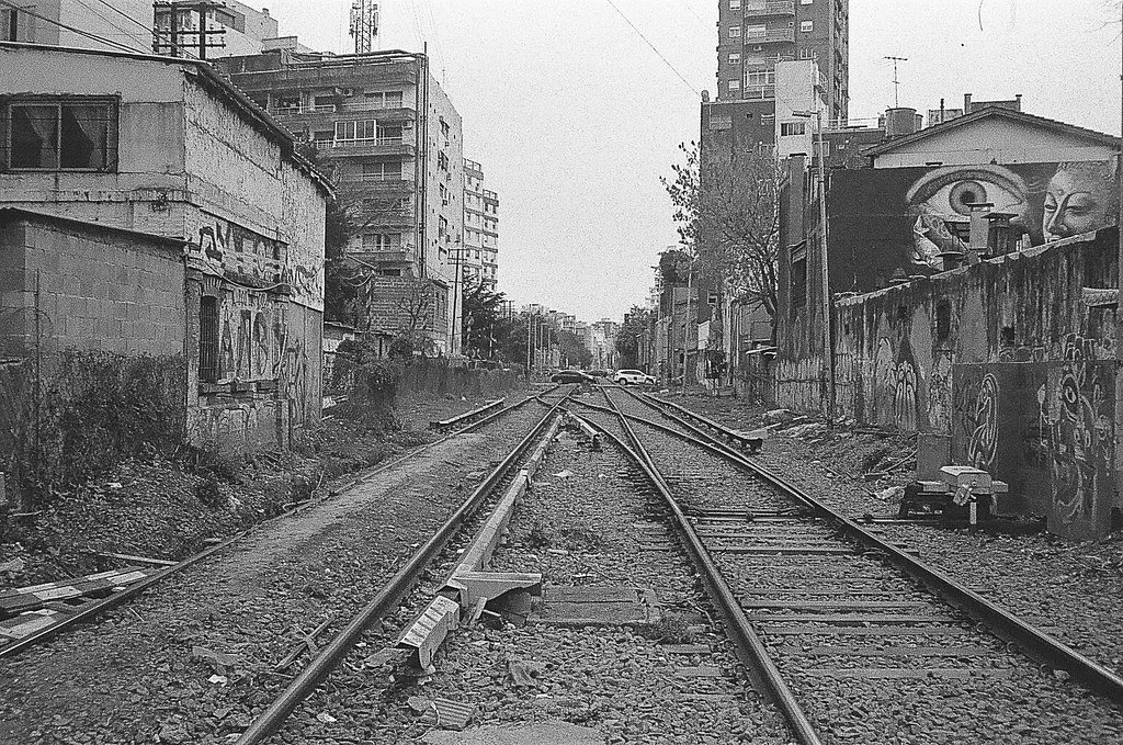 Imagem em preto e branco de via férrea no meio de uma área urbana, com prédios e paredes grafitadas ao redor.