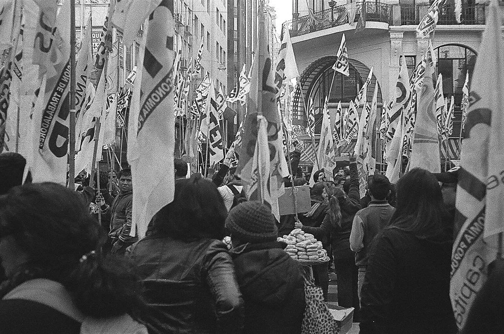 Manifestação em rua urbana com pessoas carregando bandeiras e cartazes, com edifícios ao fundo.