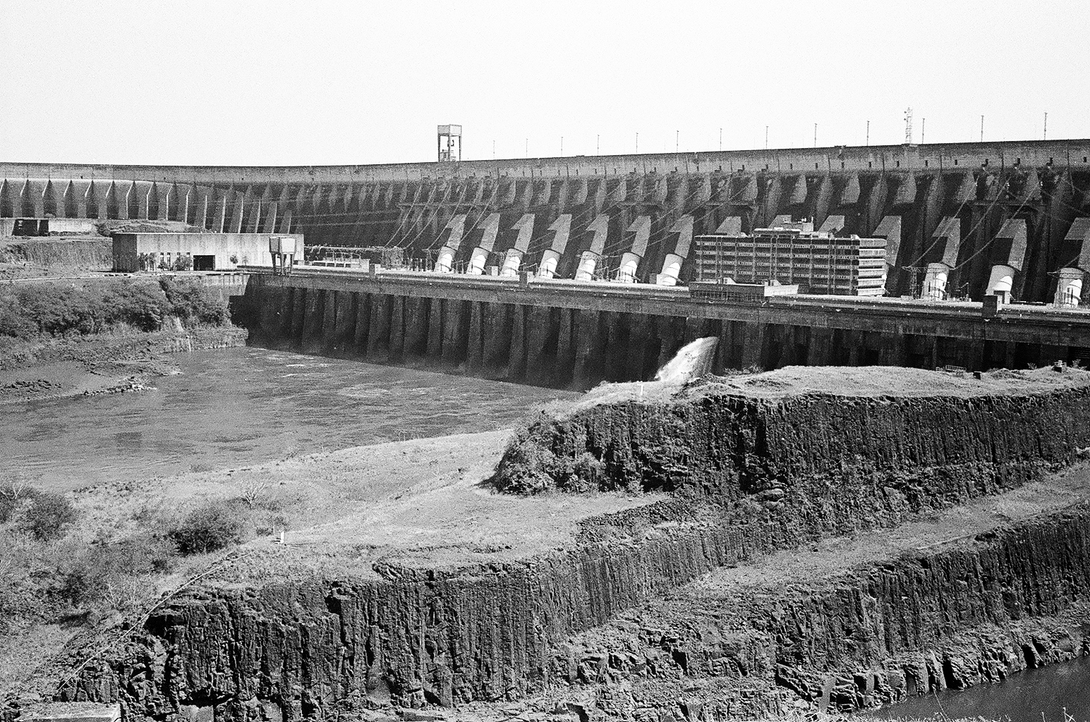 Imagem do reservatório de uma usina hidrelétrica com destaque para as barragens, comportas, torres e margens rochosas ao redor.
